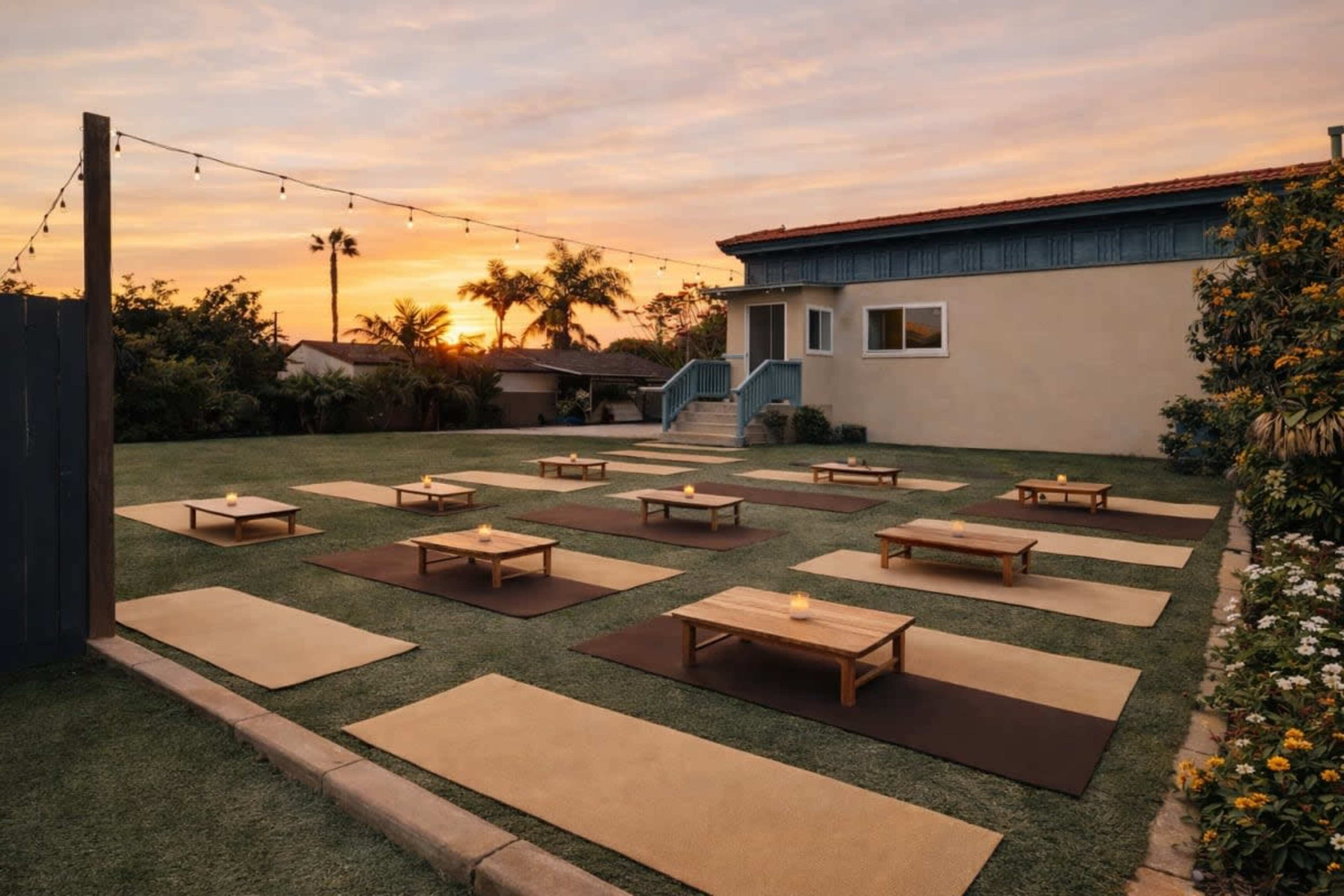 The image shows an outdoor yoga setup with several mats arranged in rows on green grass, illuminated by candles, under a sunset sky.