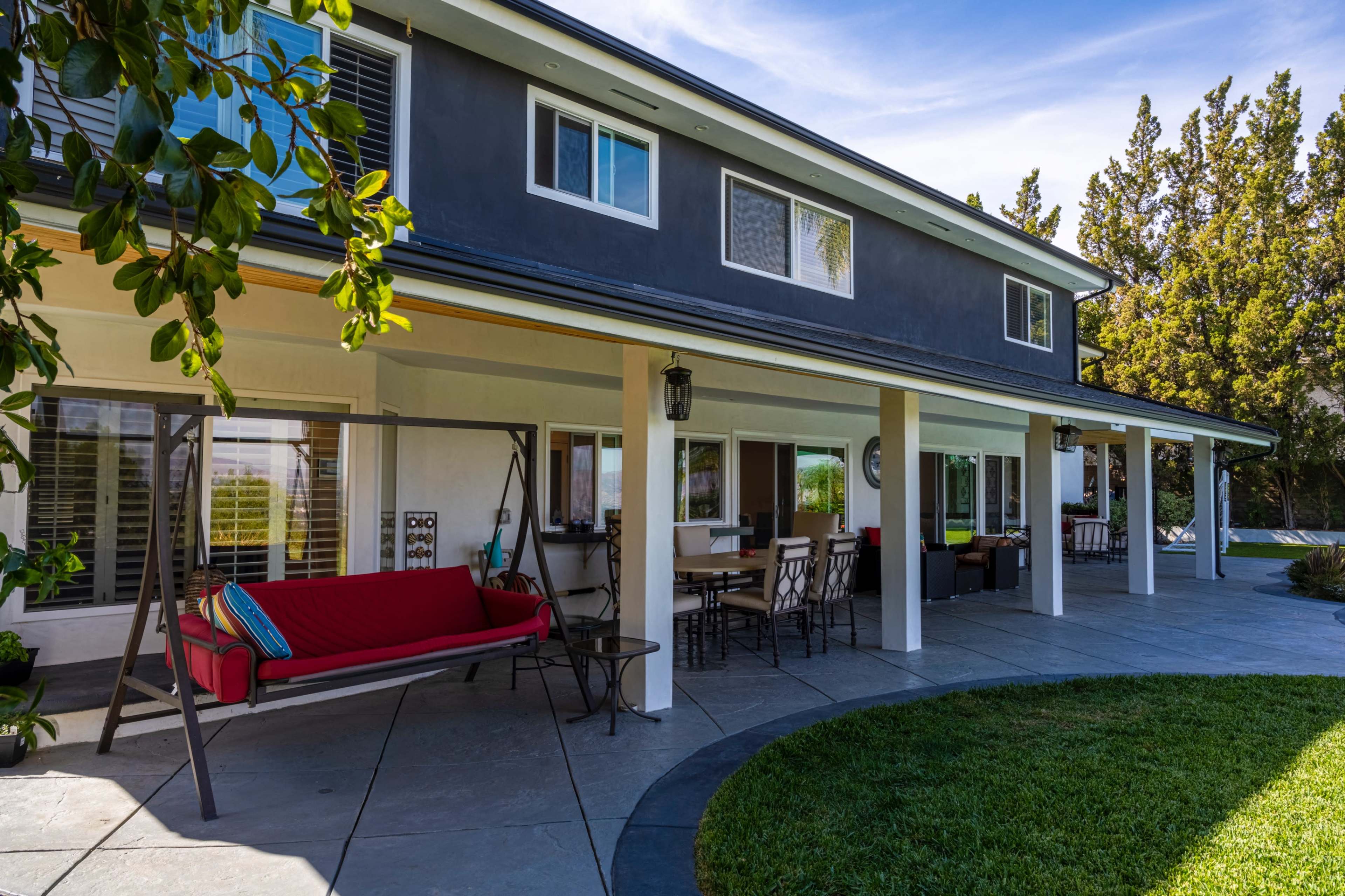 A modern two-story house with a covered patio, swing seat, and outdoor dining area surrounded by landscaped greenery.