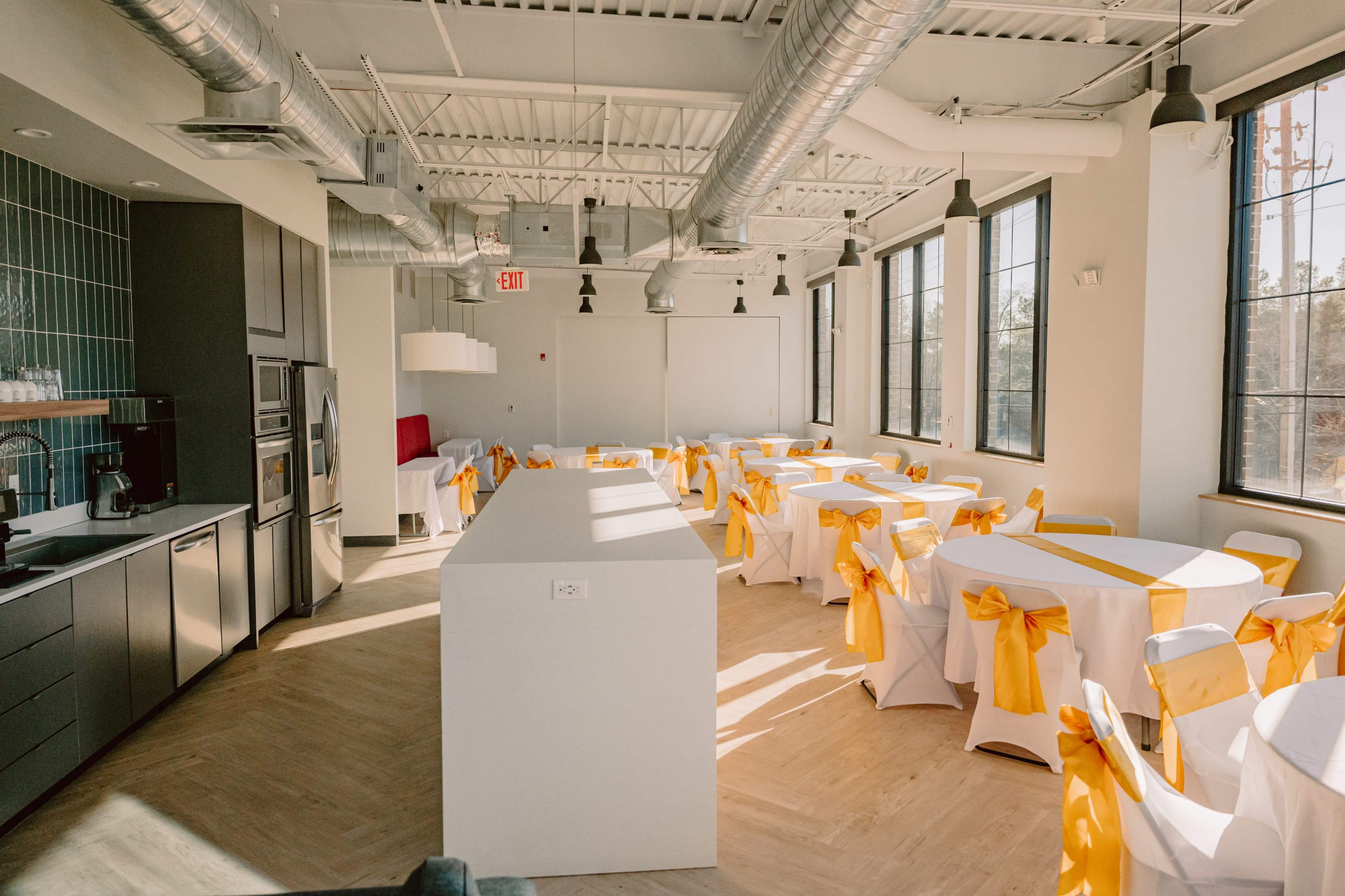 A modern event space with round tables covered in white linens and adorned with yellow satin sashes, arranged around a central white countertop.