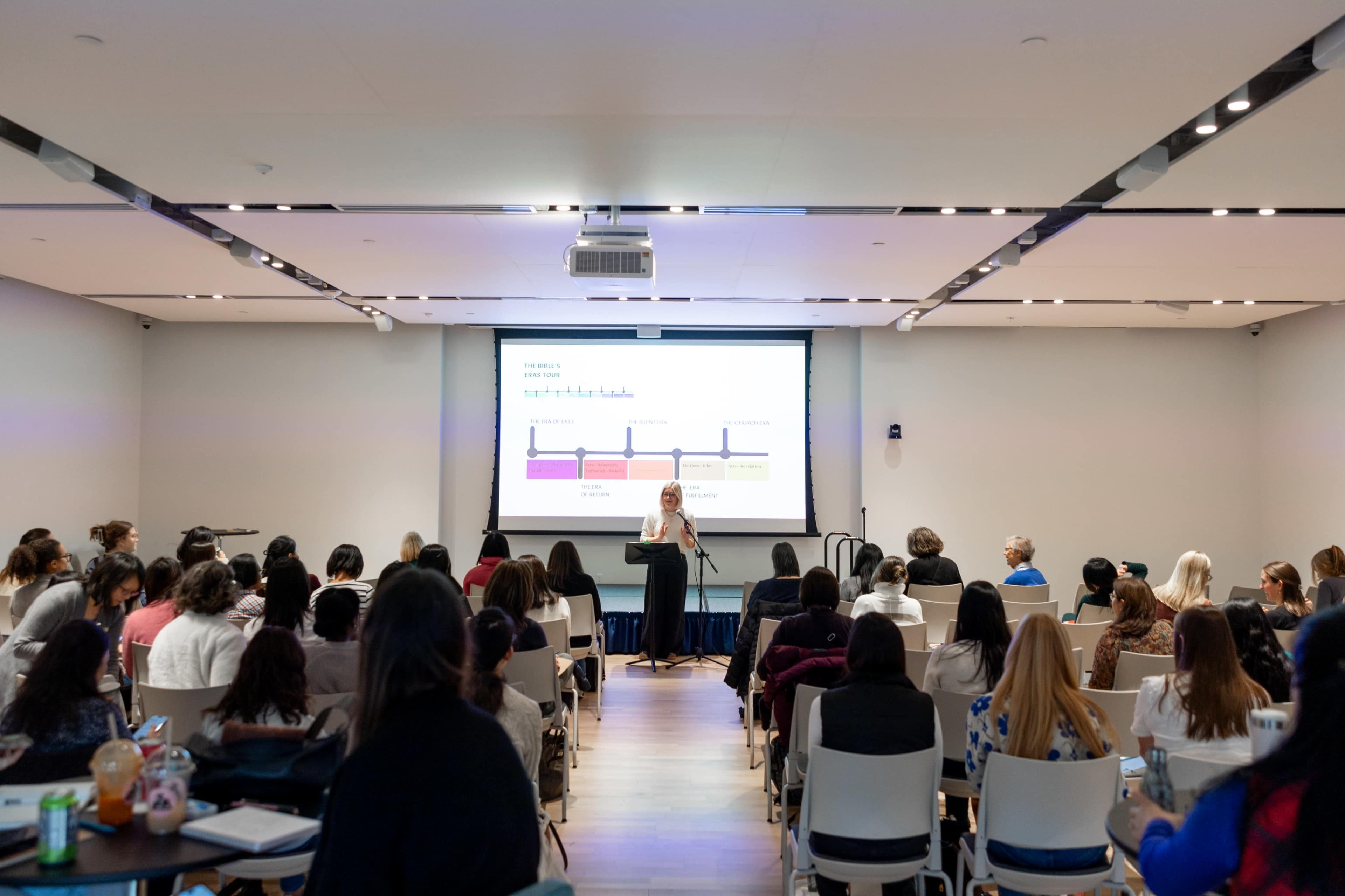 A speaker stands at the front of a conference room, presenting to an audience seated in rows while a screen displays visual aids related to the presentation.