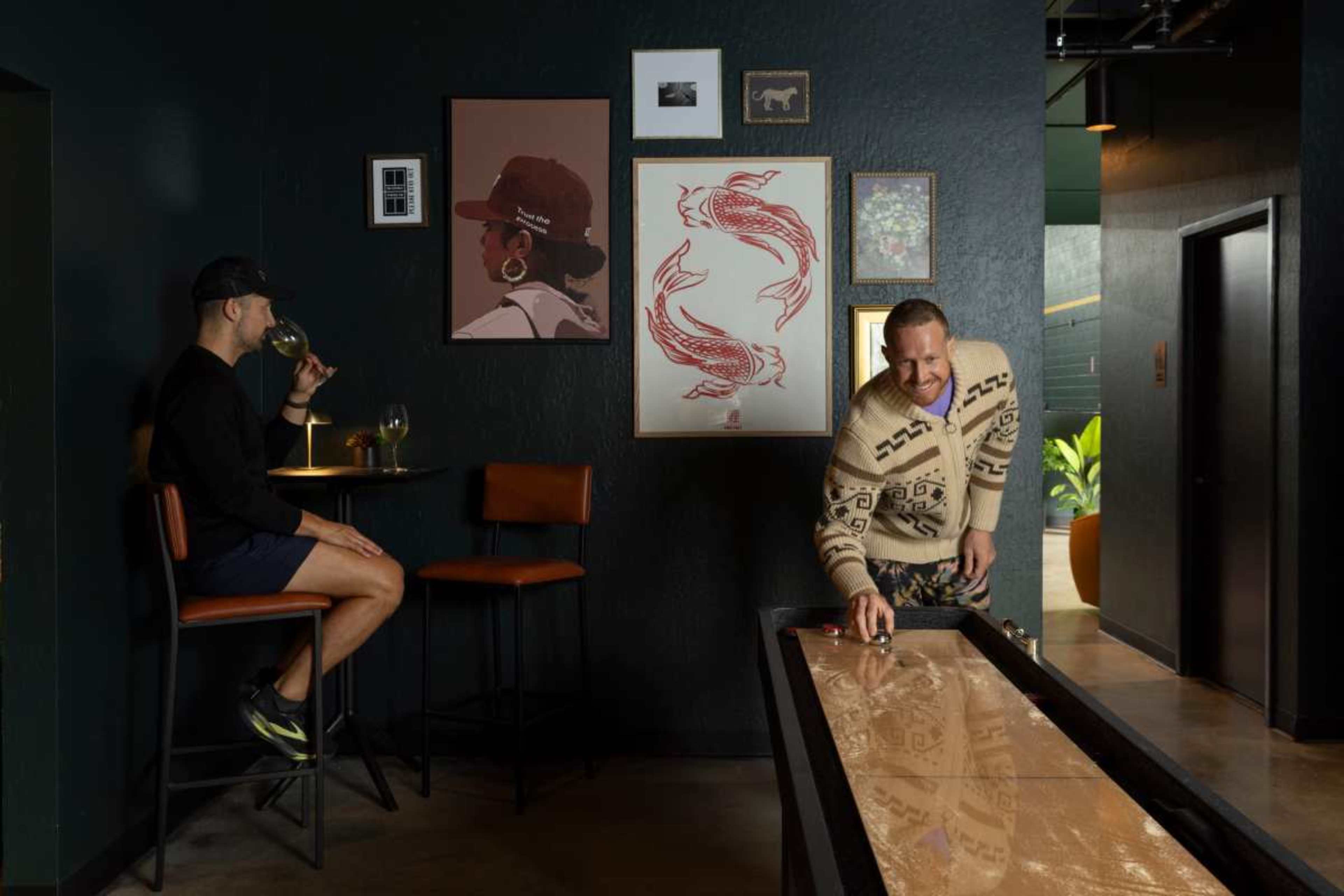 Two men are indoors at a bar; one is seated with a drink, while the other is playing a game on a wooden shuffleboard.