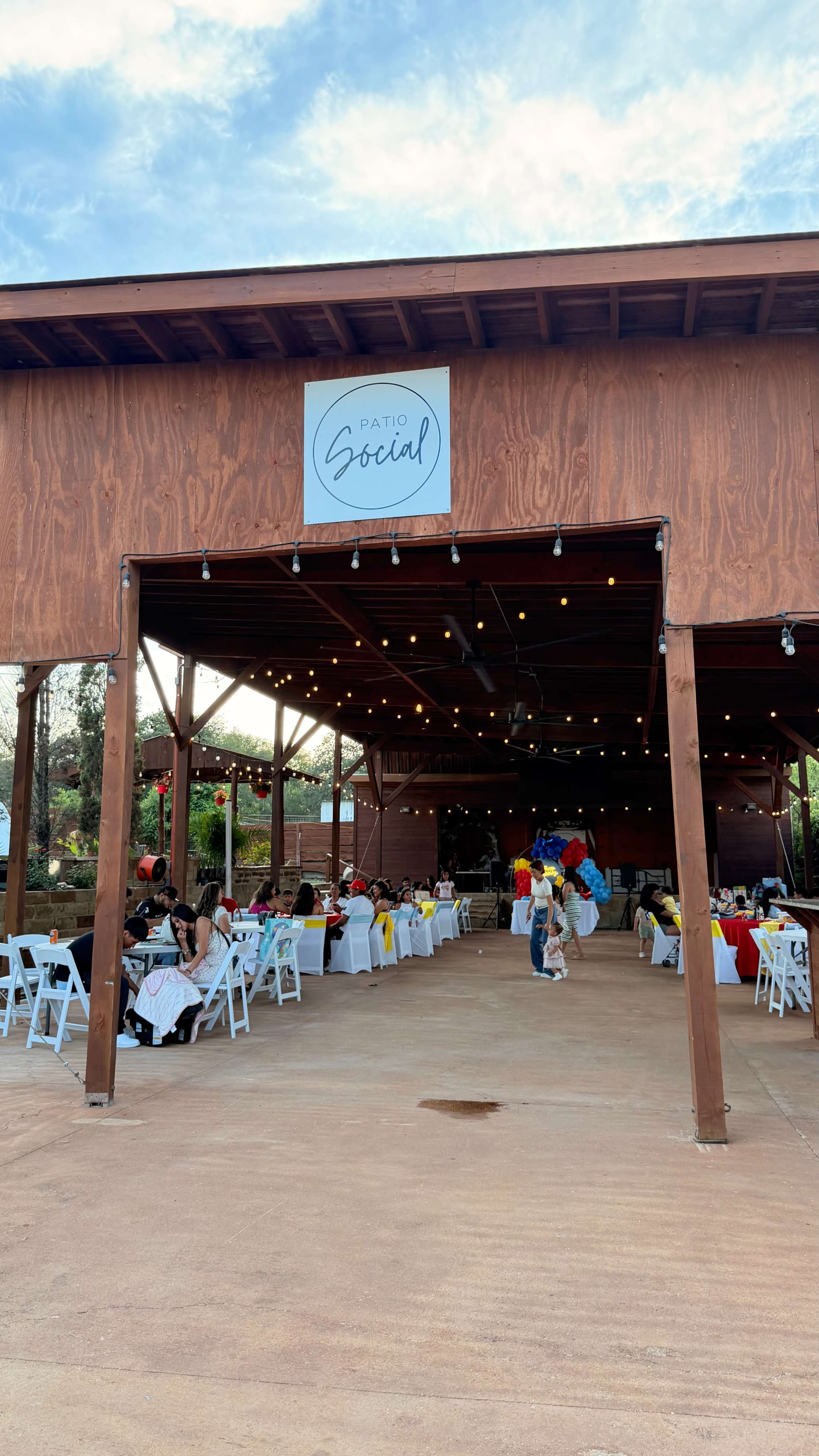 The image shows a large outdoor event space featuring tables set with colorful tablecloths, under a wooden structure with a sign that reads "Patio Social."