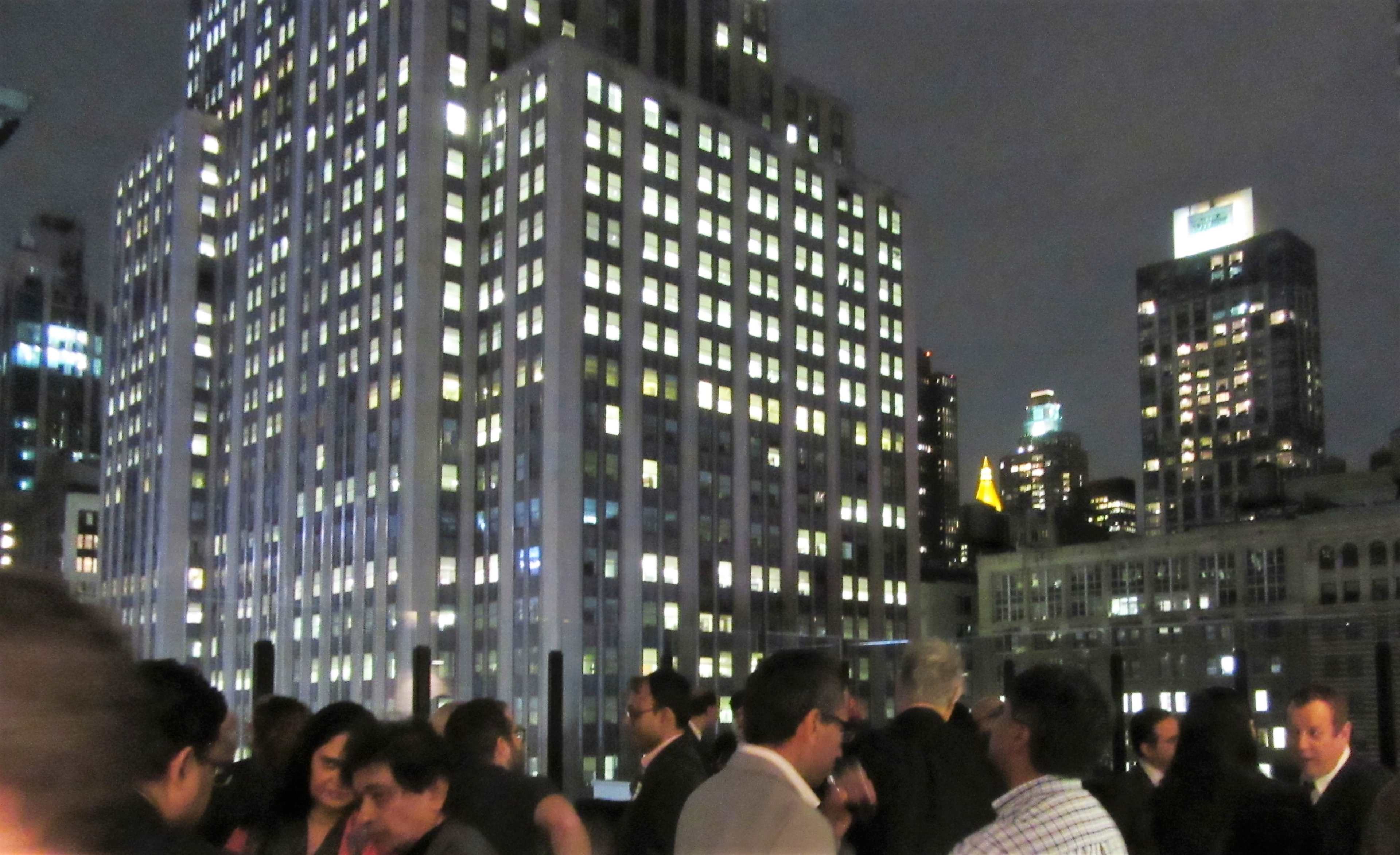 A bustling rooftop gathering at night, illuminated by city lights and surrounded by tall skyscrapers.