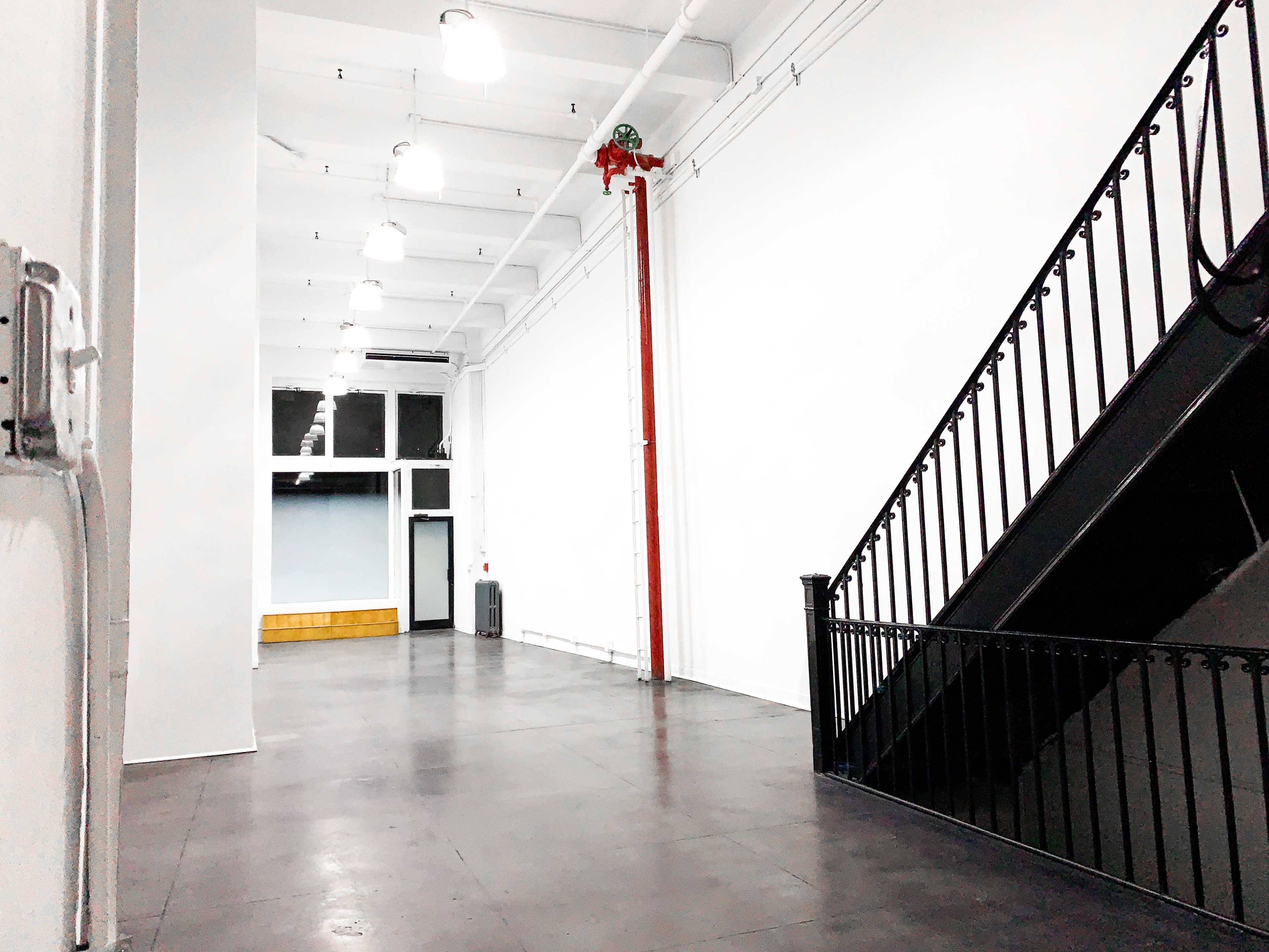An empty industrial-style interior with white walls, a staircase on one side, and large windows allowing light to enter.