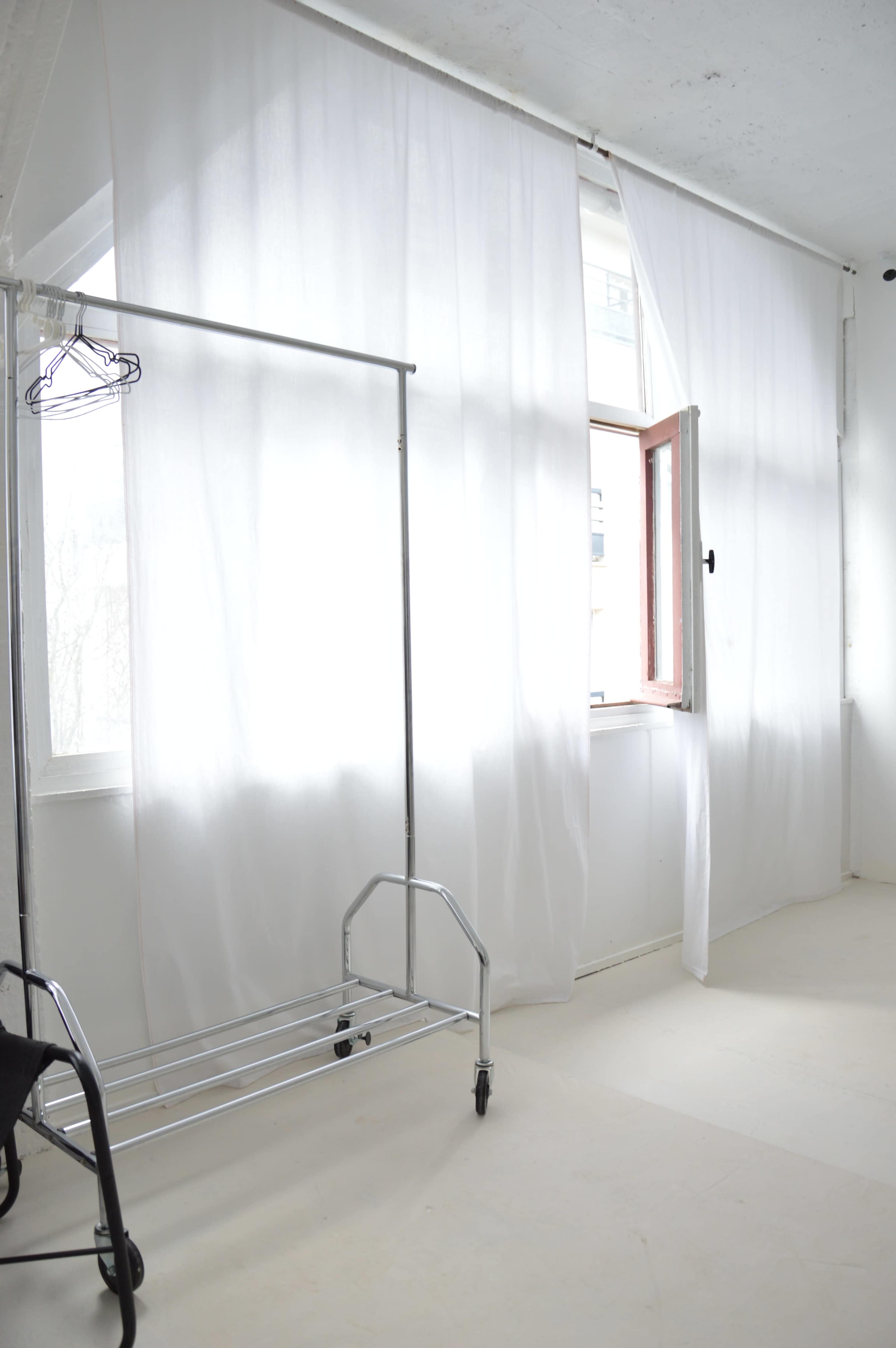 A lightweight clothes rack stands against a wall beside a window covered with sheer white curtains.