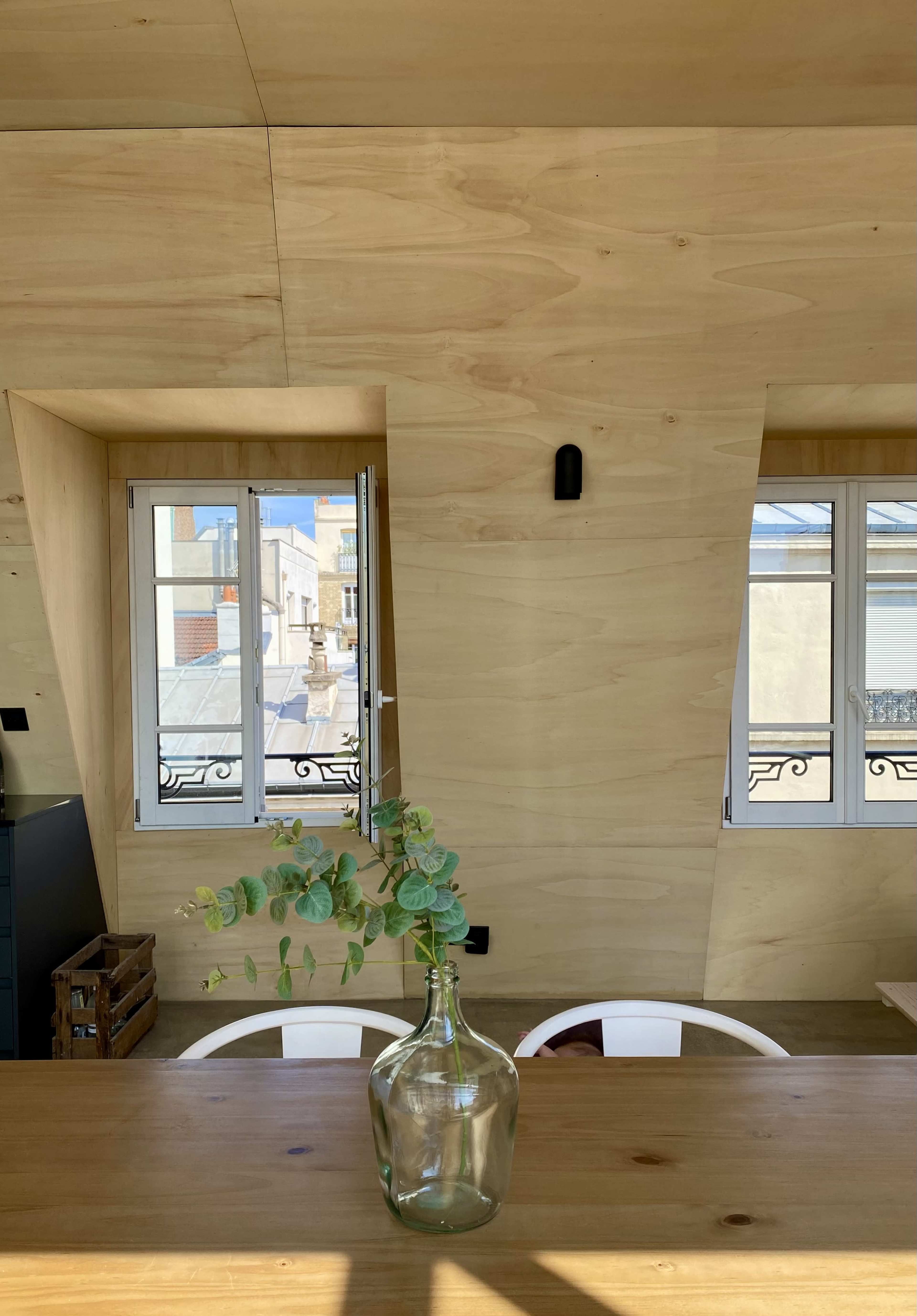 A minimalist wooden interior with a glass vase containing eucalyptus placed on a table, and two windows revealing a view of rooftops outside.