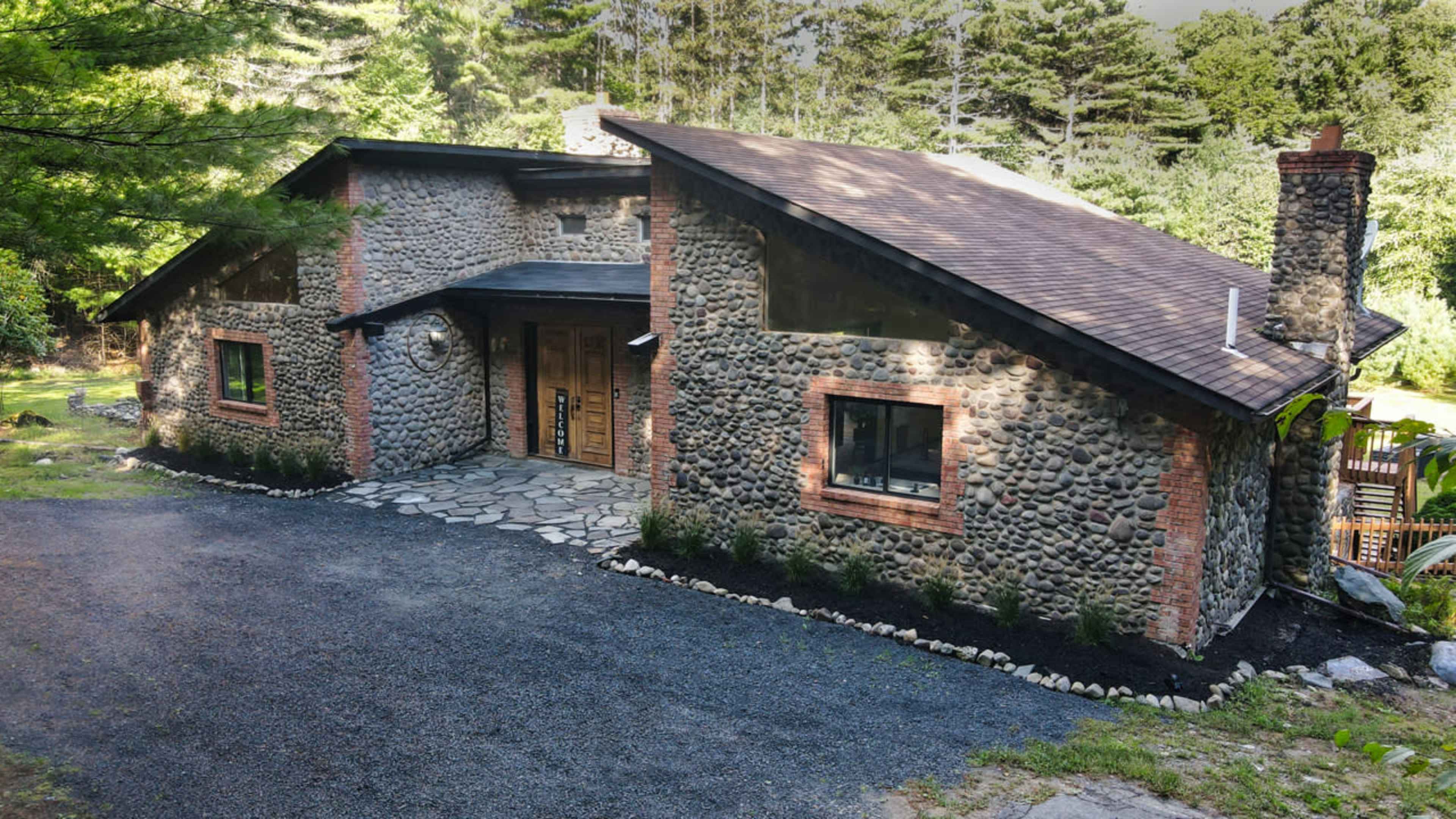 A stone and brick house with a sloped roof is set in a wooded area, featuring a landscaped pathway and outdoor seating.