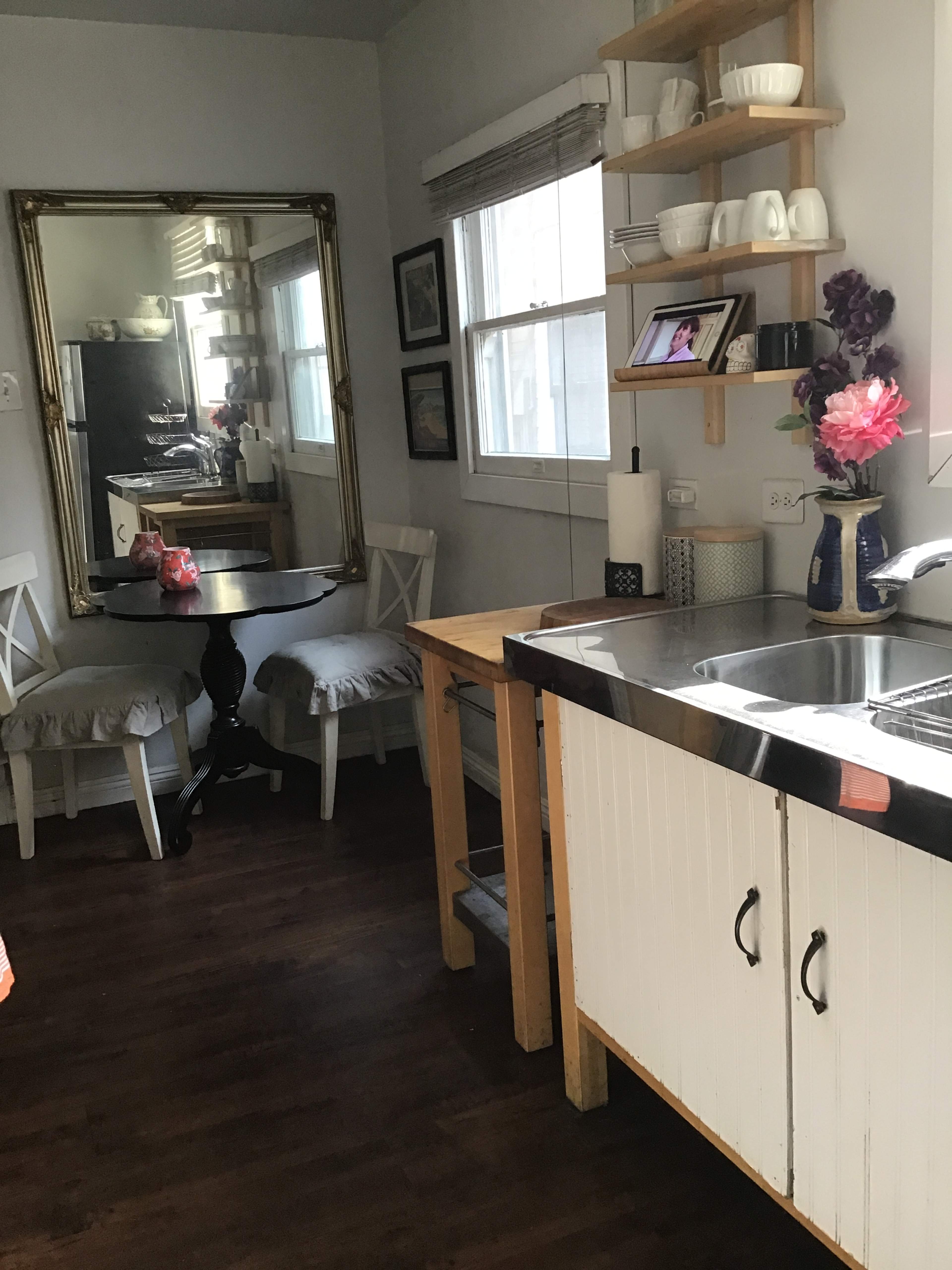 A small kitchen area featuring a table, two chairs, a mirrored wall, and a stainless steel sink with wooden cabinets.