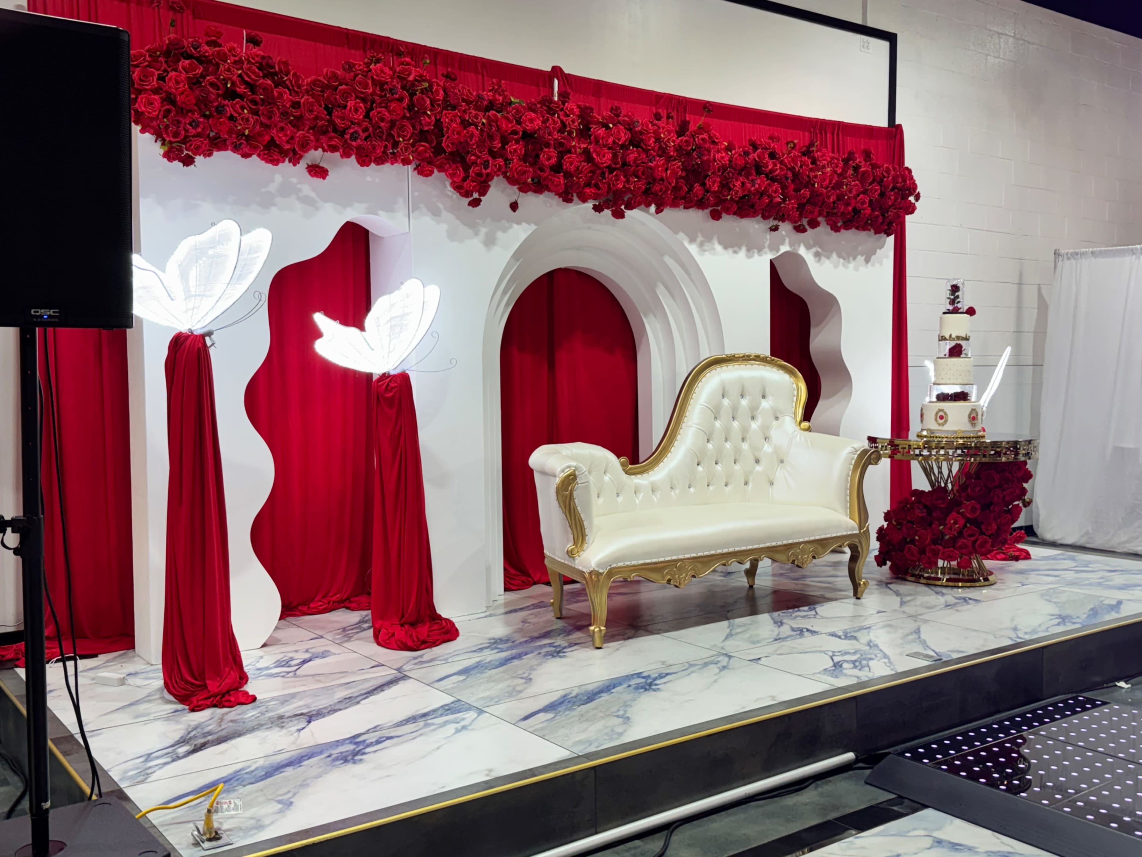 The image displays a decorated event space featuring a white sofa with gold accents, flanked by floral arrangements, a tiered cake, and a backdrop of red curtains and roses.
