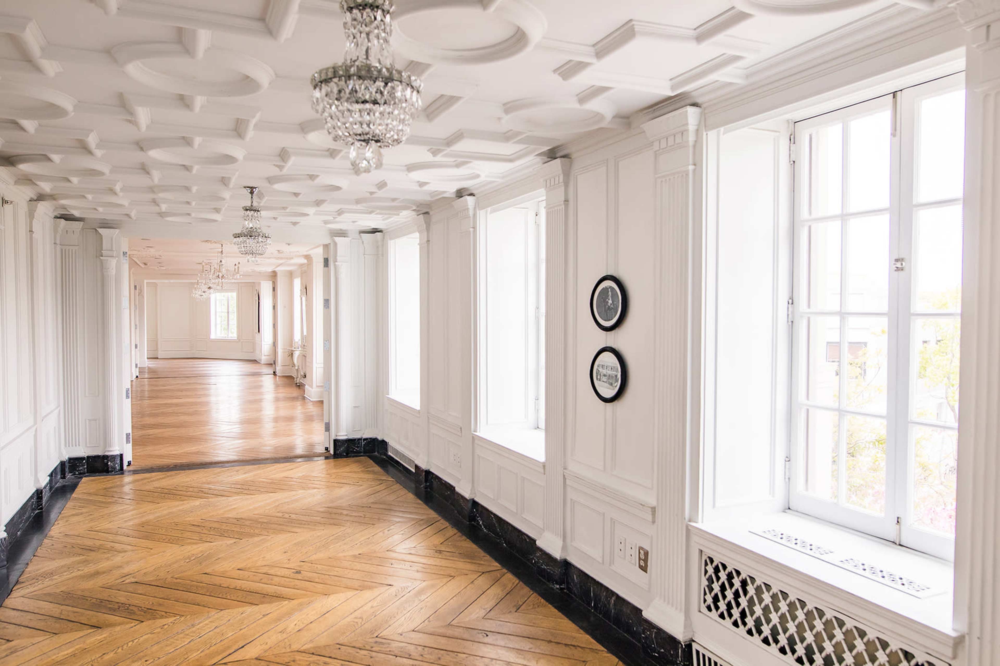 The image shows a corridor with white walls, ornate ceilings, and large windows, featuring a wooden floor with a herringbone pattern.