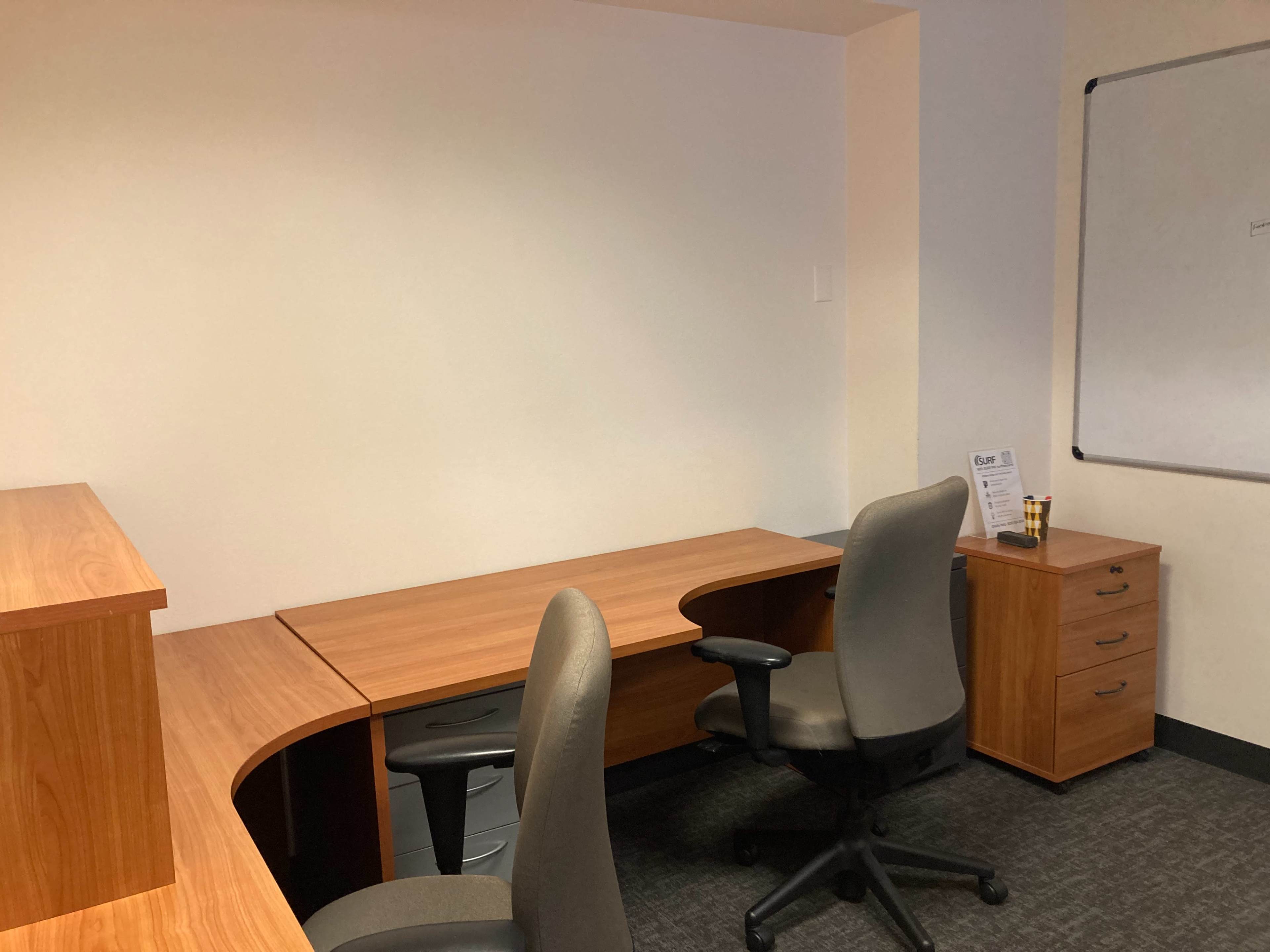 The image shows a minimalistic office space with two wooden desks, a filing cabinet, and a whiteboard on the wall.