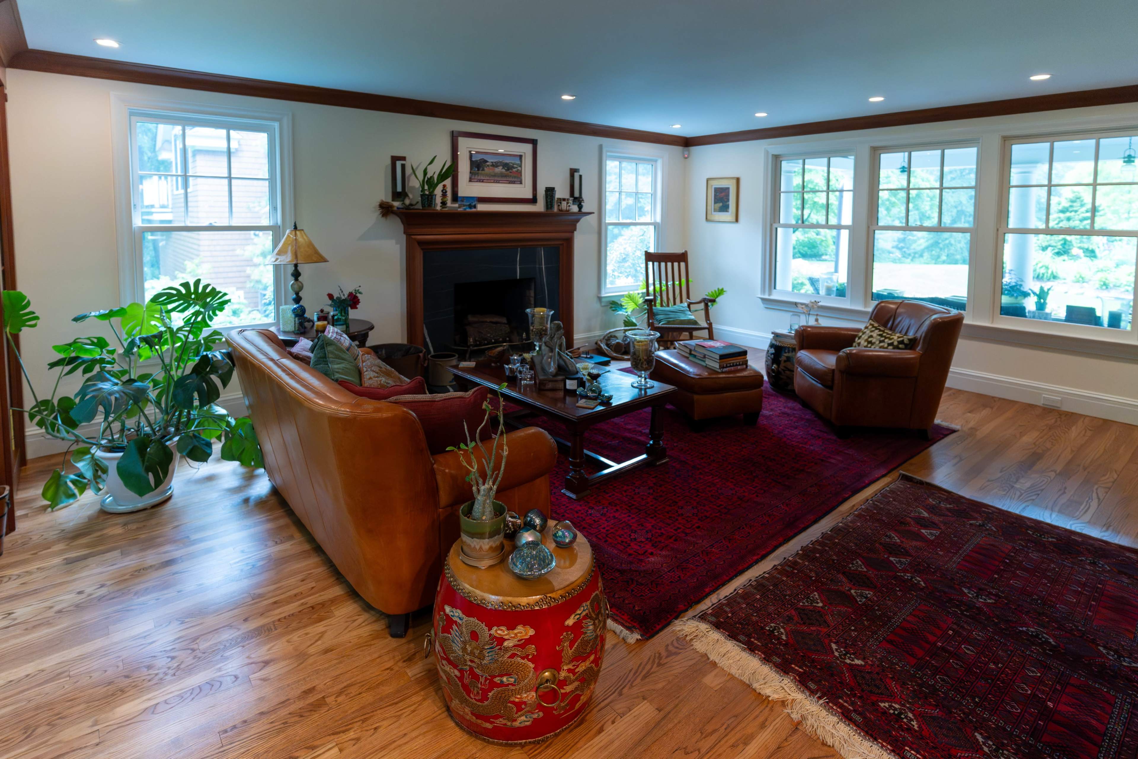 The image shows a living room with a brown leather sofa, two armchairs, a coffee table, and large windows allowing natural light to enter.