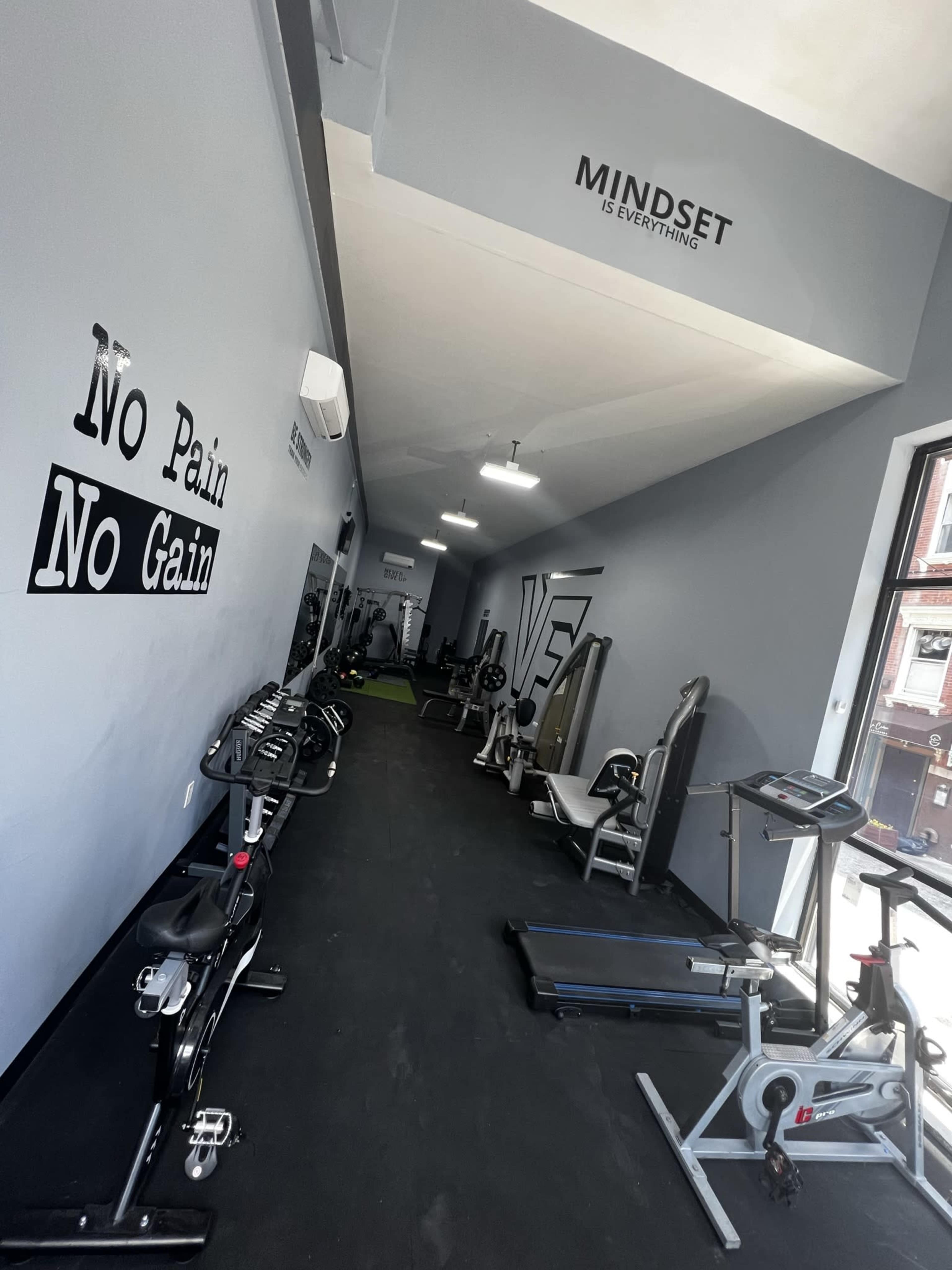 The image shows a narrow fitness studio with exercise equipment lined along the walls and motivational phrases on the gray walls.