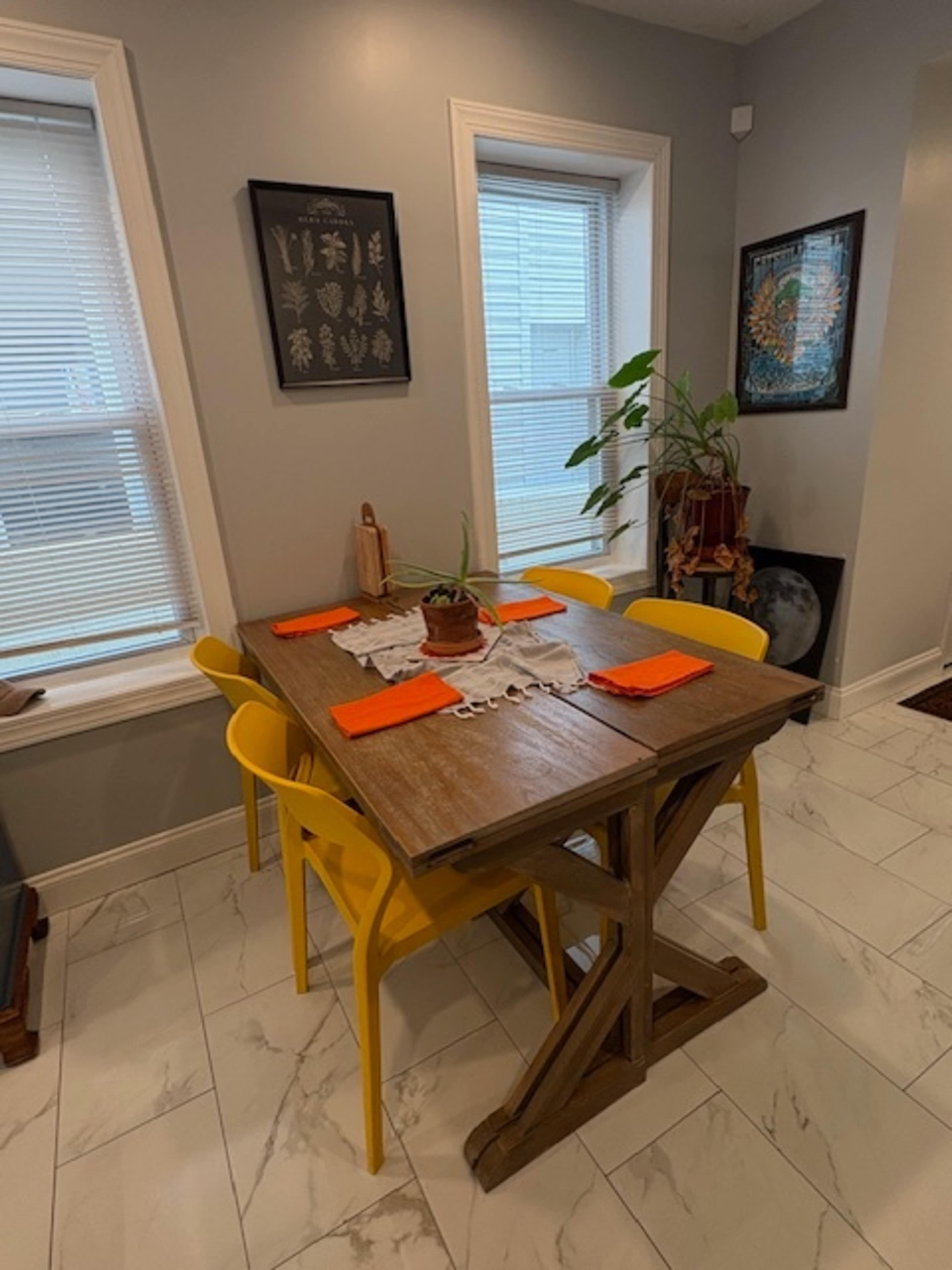 A wooden dining table with orange placemats and yellow chairs is set in a well-lit dining area with windows and decorative plants.