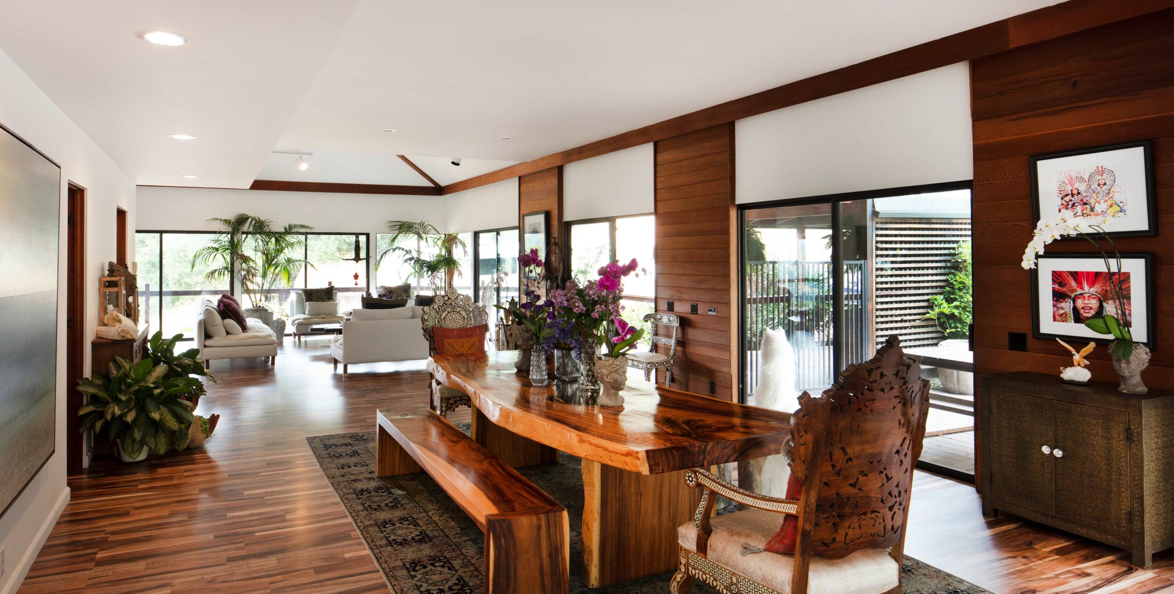 A spacious modern living area featuring a wooden dining table and decorative plants, with large windows allowing natural light to fill the room.