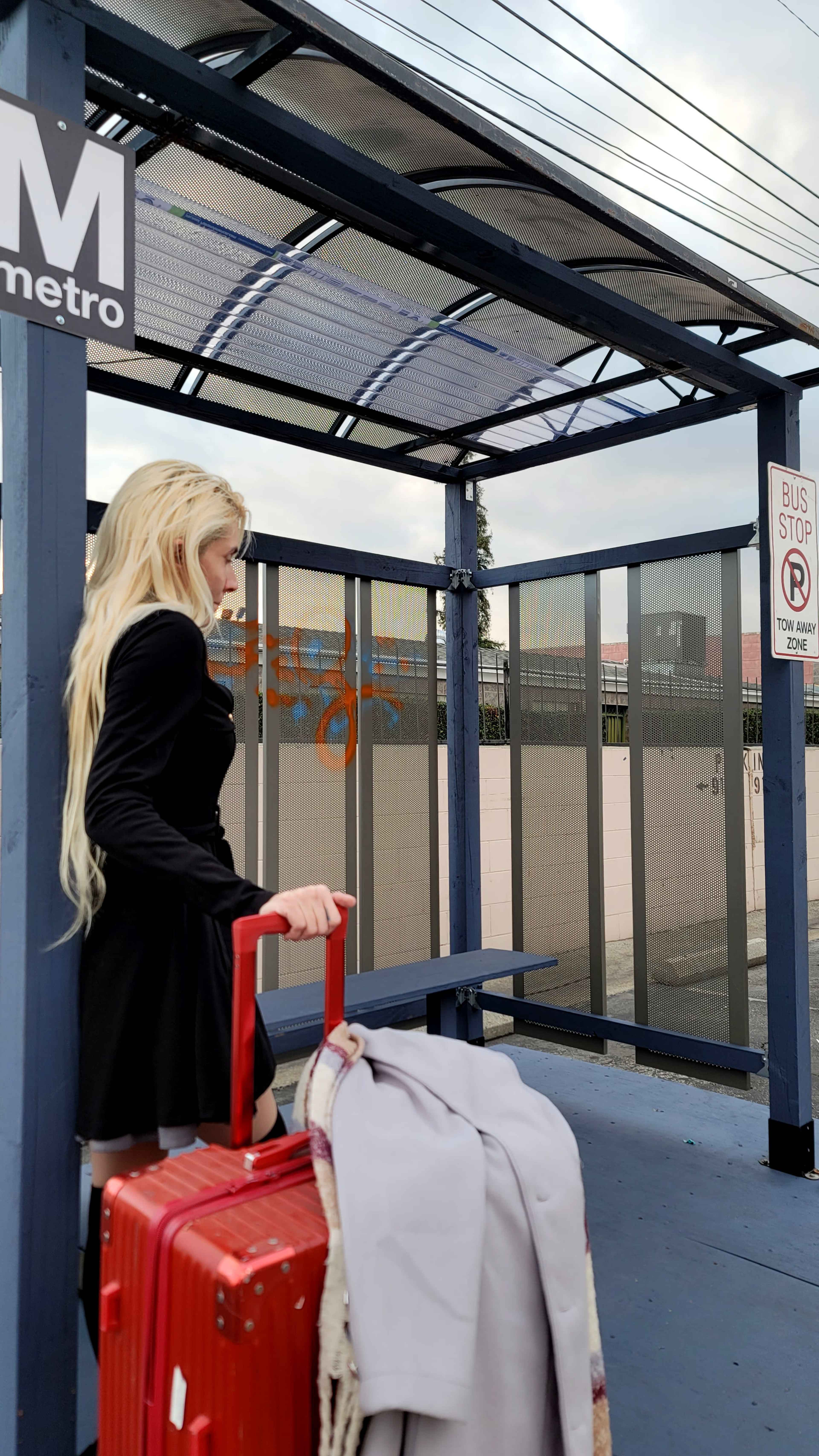 A person with long, blonde hair stands at a bus stop, holding a red suitcase and a light-colored garment draped over their arm.