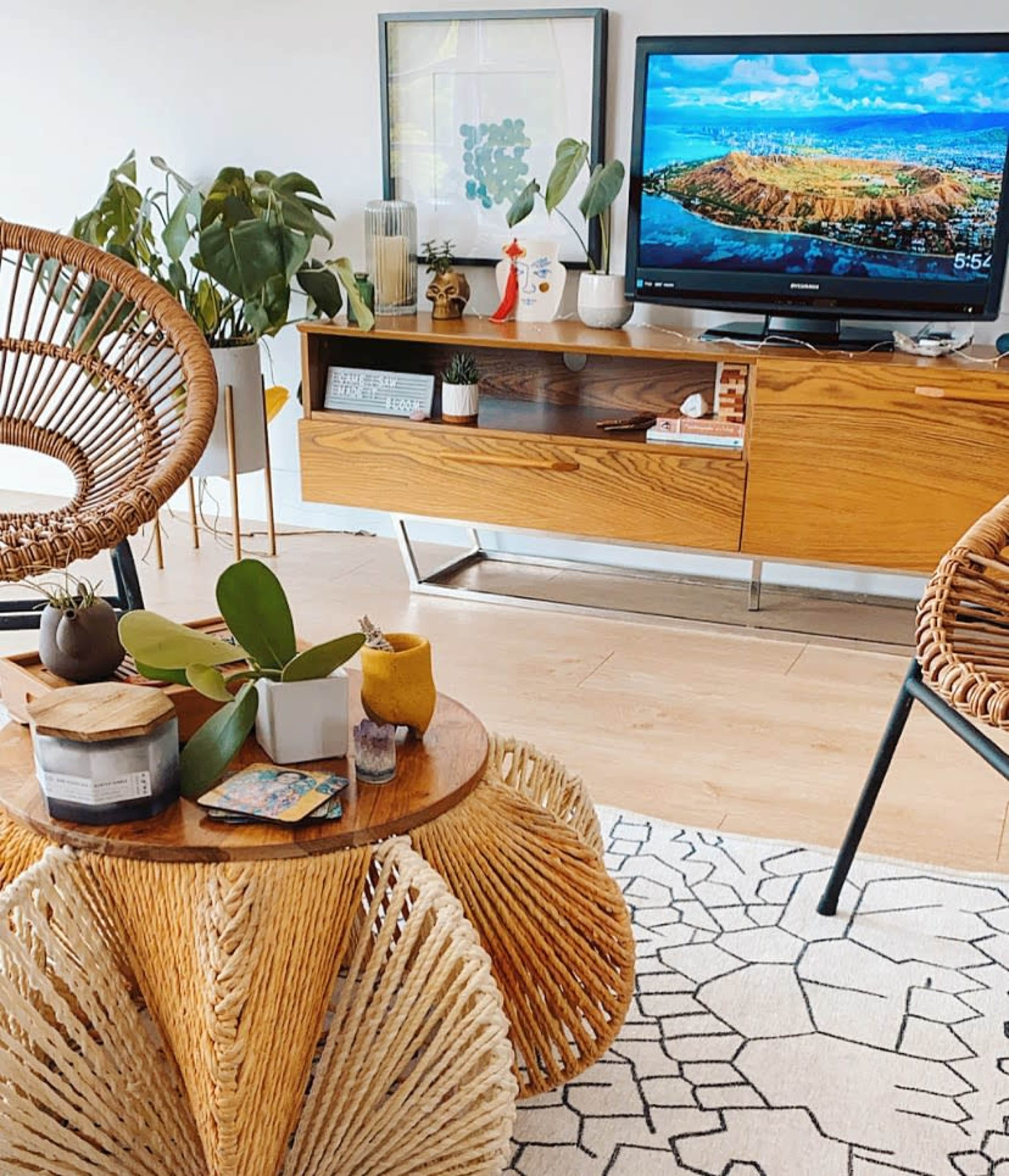 A living room area with wicker chairs, a round wooden coffee table, a television displaying a scenic view, and a wooden sideboard with plants and decorative items.