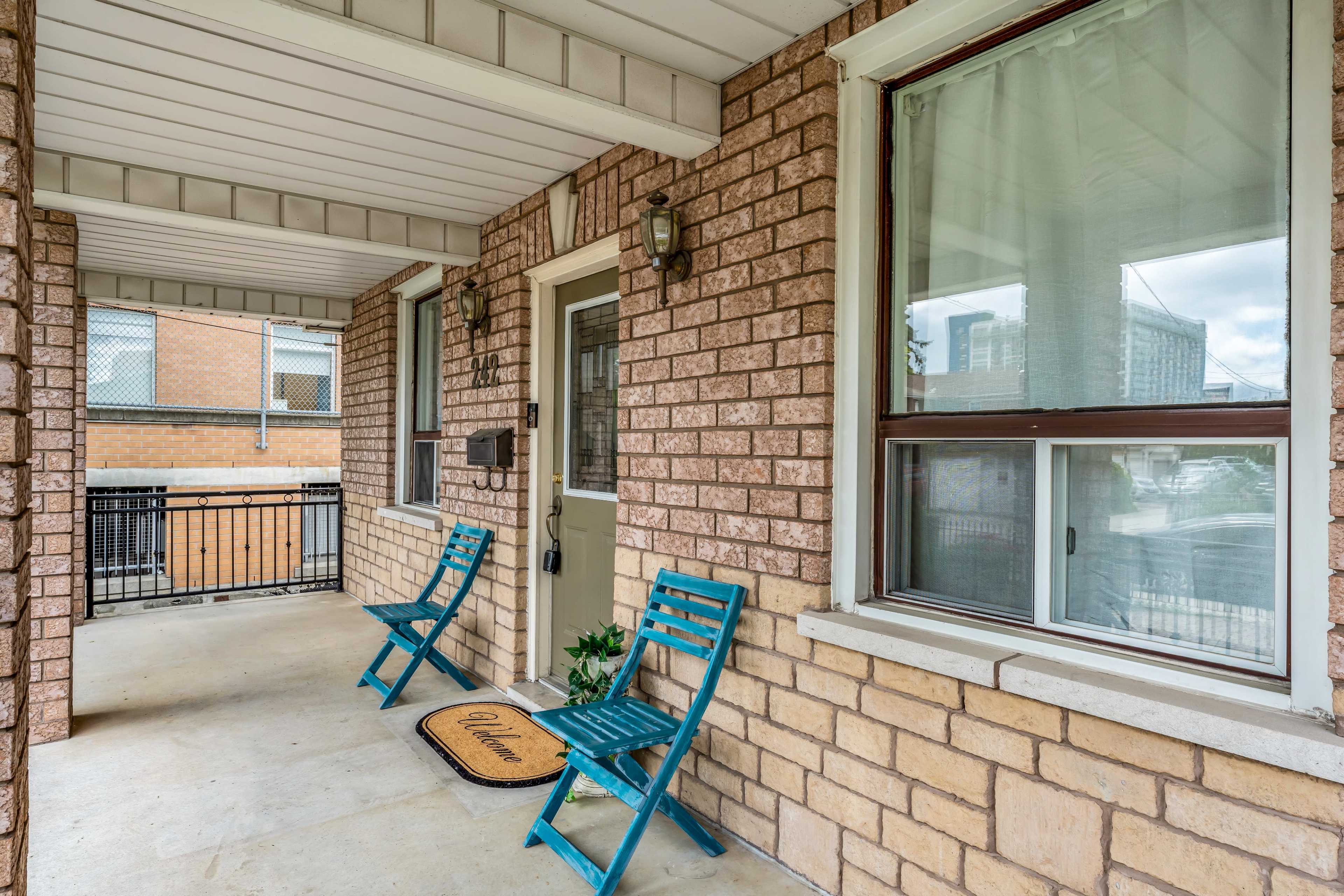 The image shows a porch area featuring a door with a welcome mat and two blue chairs positioned nearby.