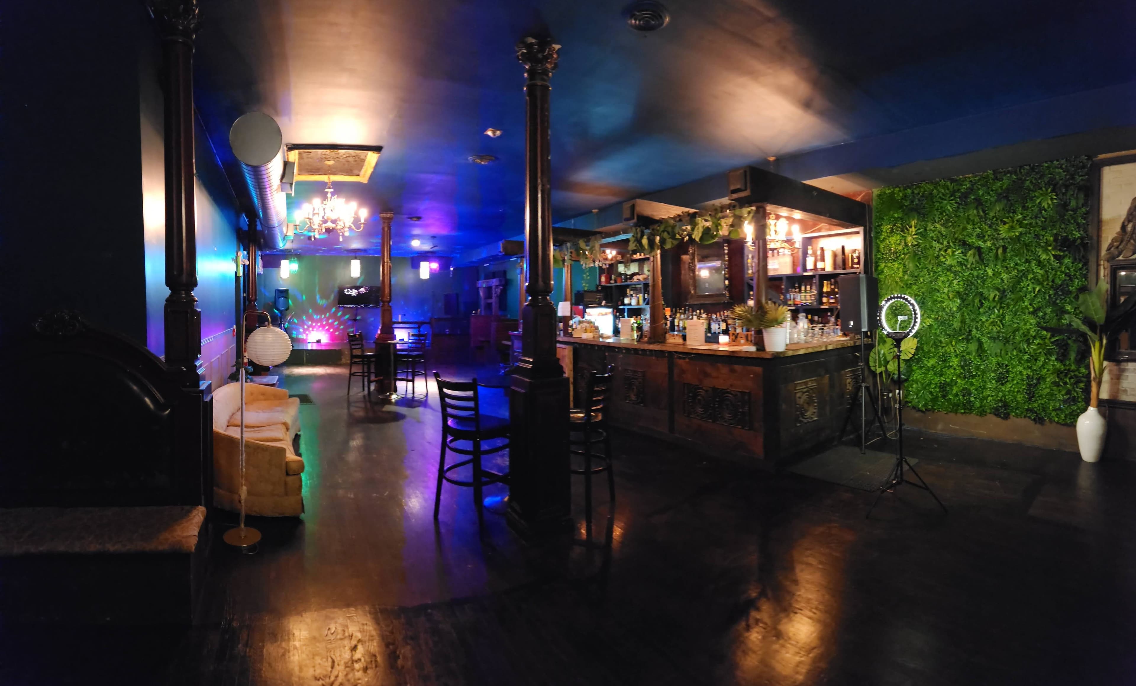 An indoor bar area with a wooden bar, lounge seating, a green wall, and dim lighting accented by colorful lights.