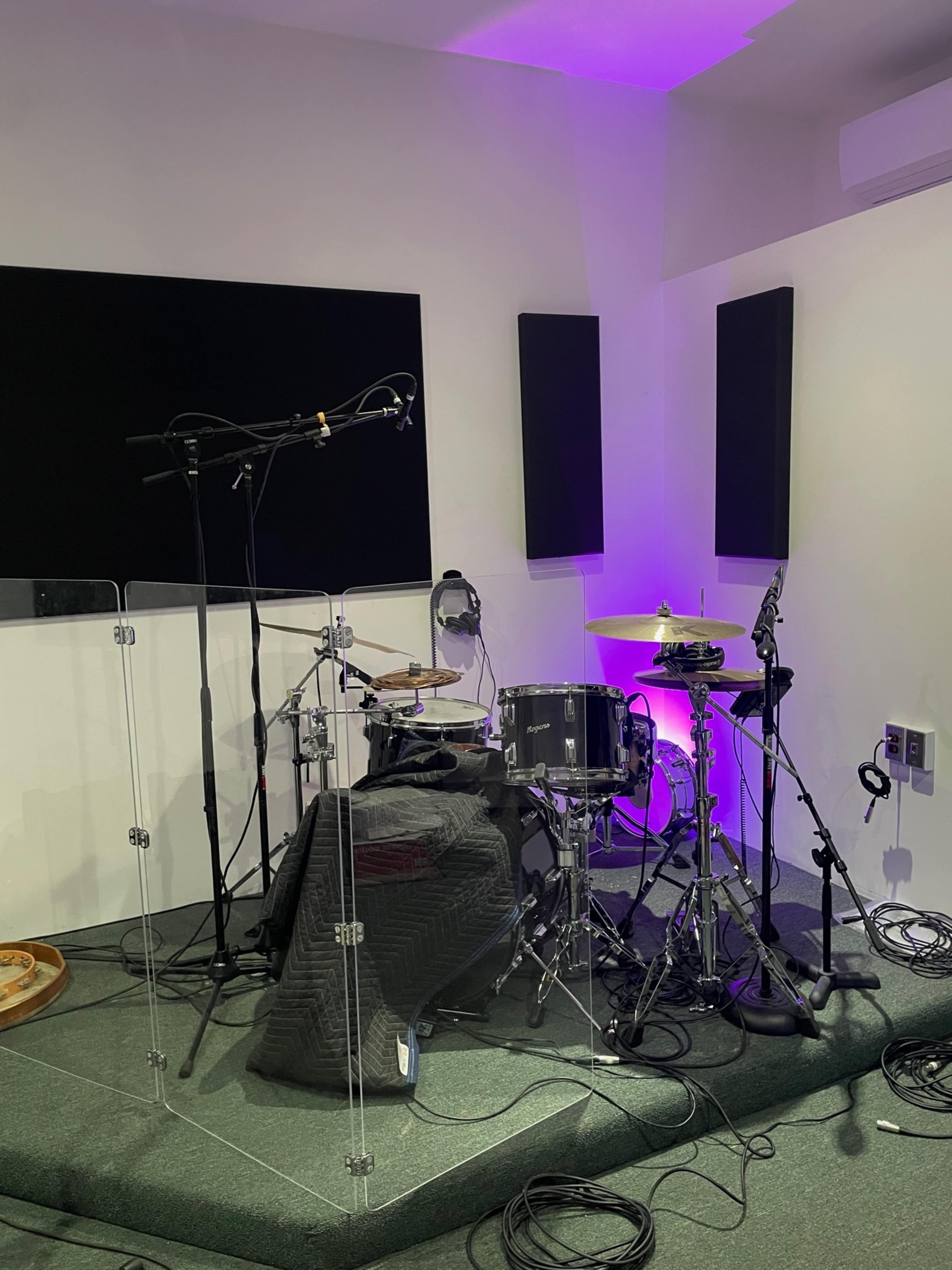 A drum set enclosed by transparent acrylic panels in a soundproof studio room with purple lighting and audio equipment.