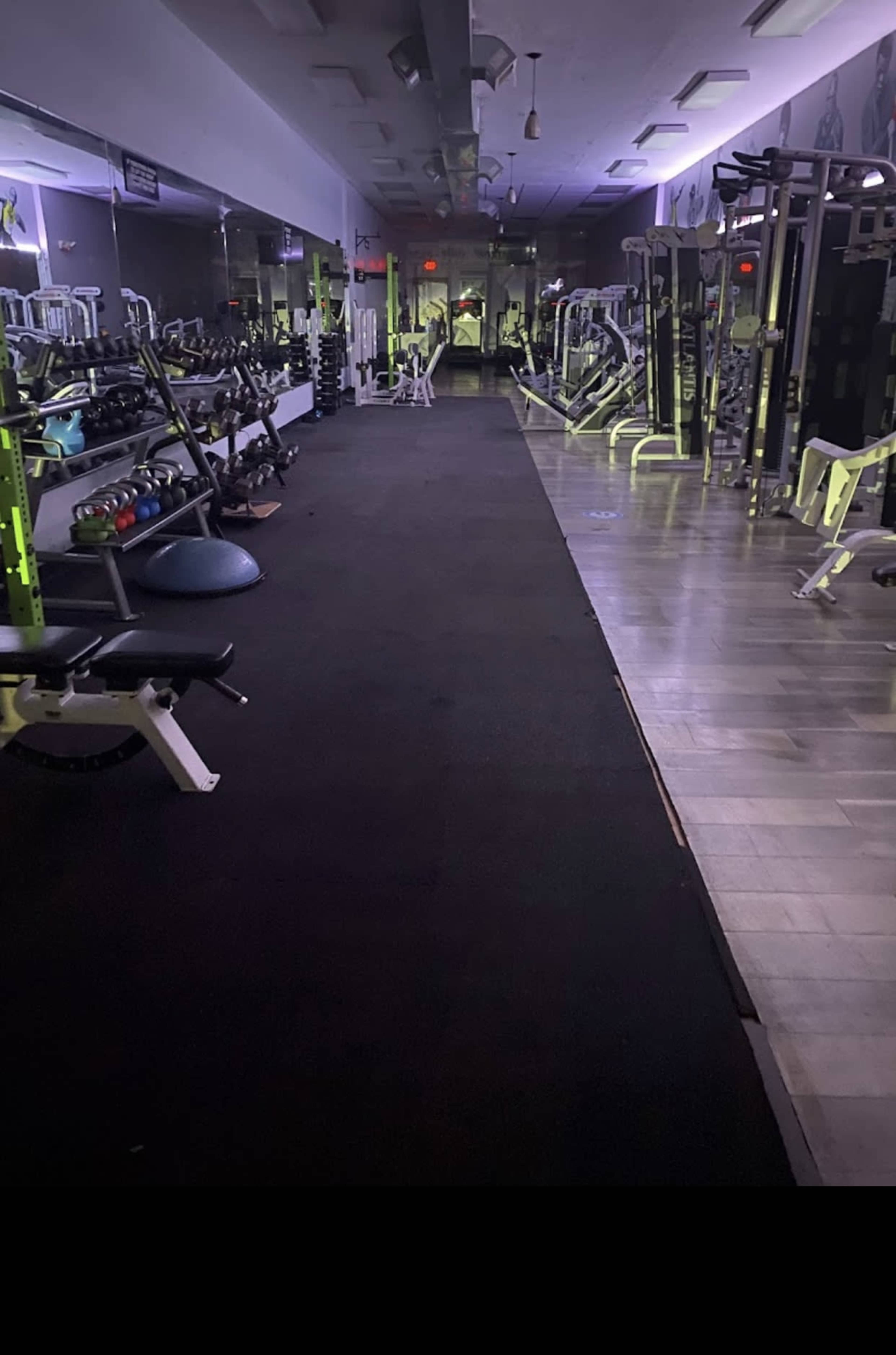 A dimly lit gym with various workout machines and free weights arranged along both sides of a long, carpeted area.