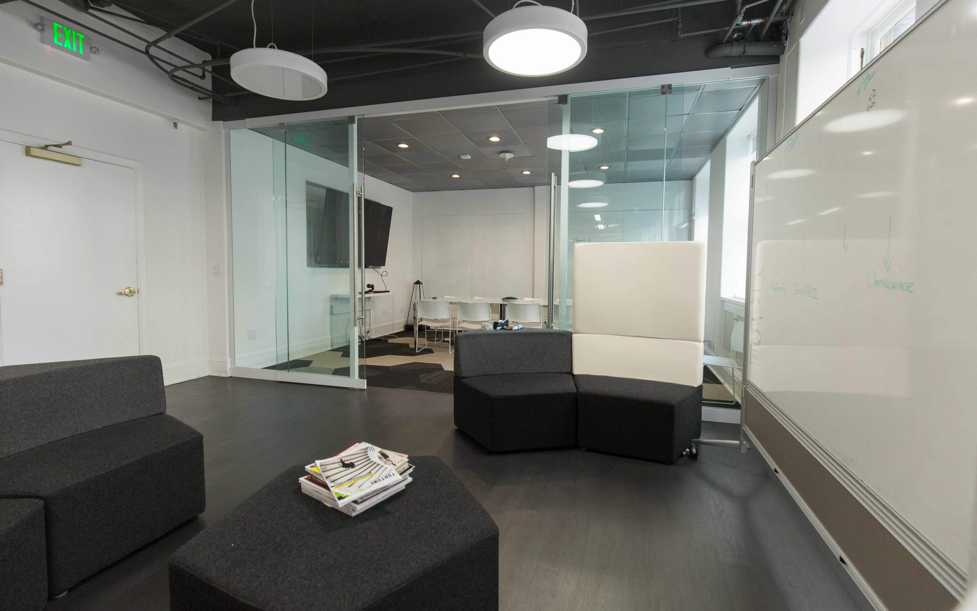 A modern office space featuring a seating area with black modular furniture, a glass-walled meeting room in the background, and a whiteboard on the right.