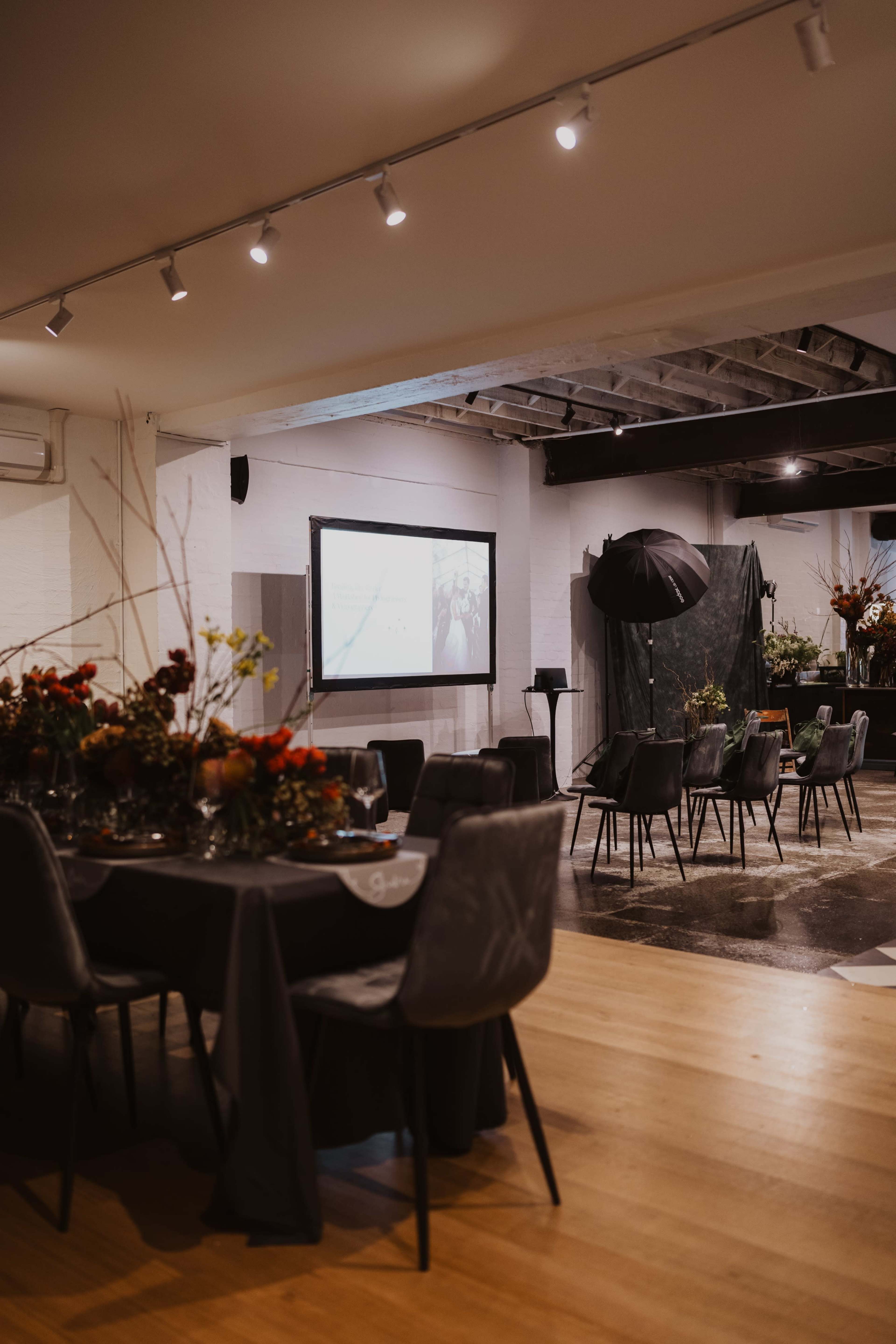 A spacious event venue features tables set for dining, a large screen for presentations, and decorative floral arrangements.