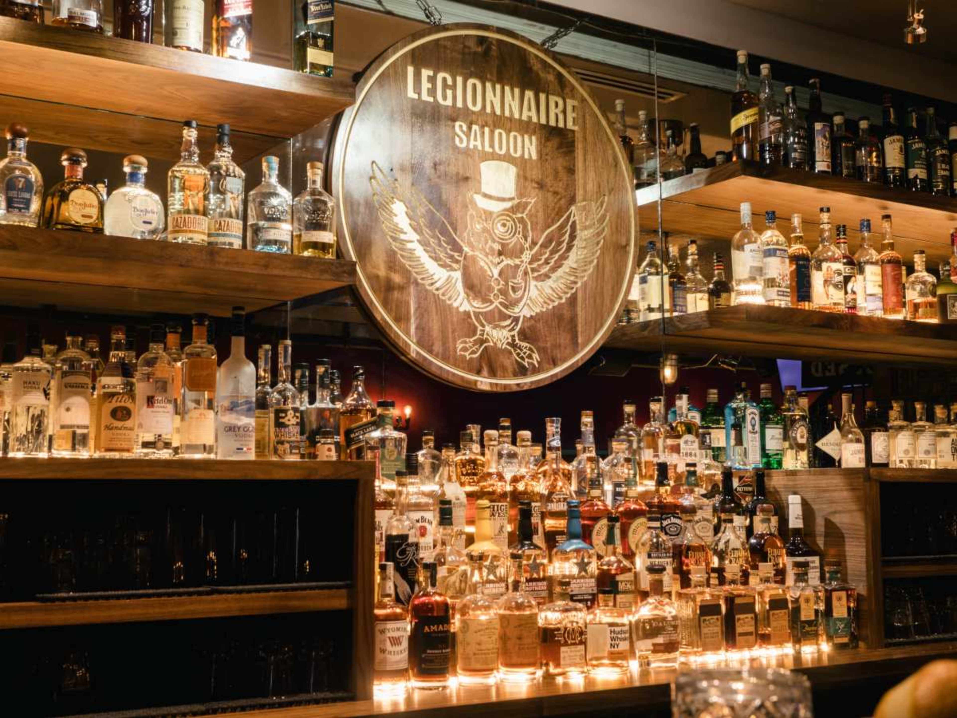 The image shows a well-stocked bar with shelves filled with various bottles of liquor and a wooden sign labeled "Legionnaire Saloon" prominently displayed at the center.