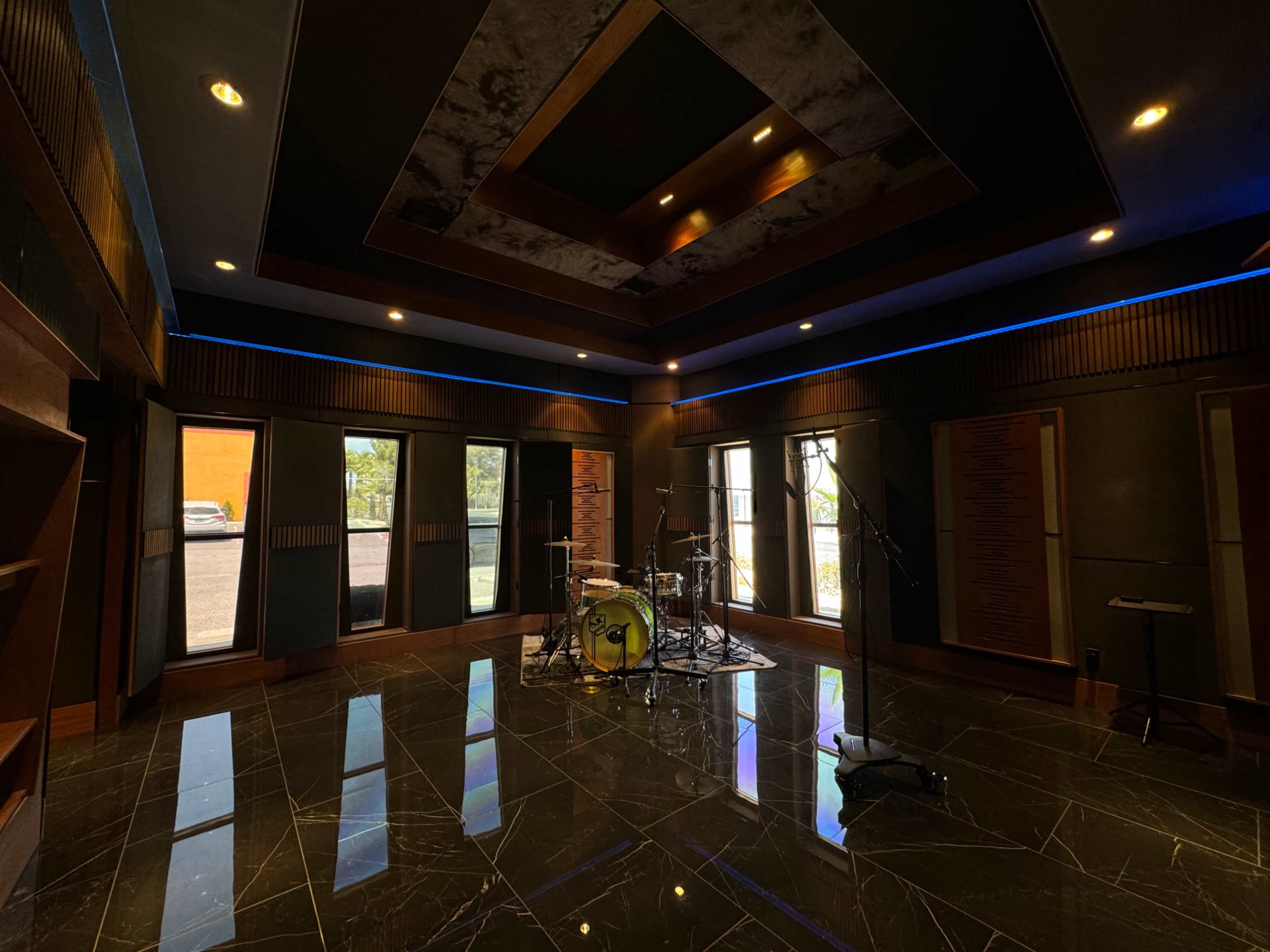 A music studio with a drum set placed on a polished black floor, surrounded by large windows and acoustic paneling on the walls.