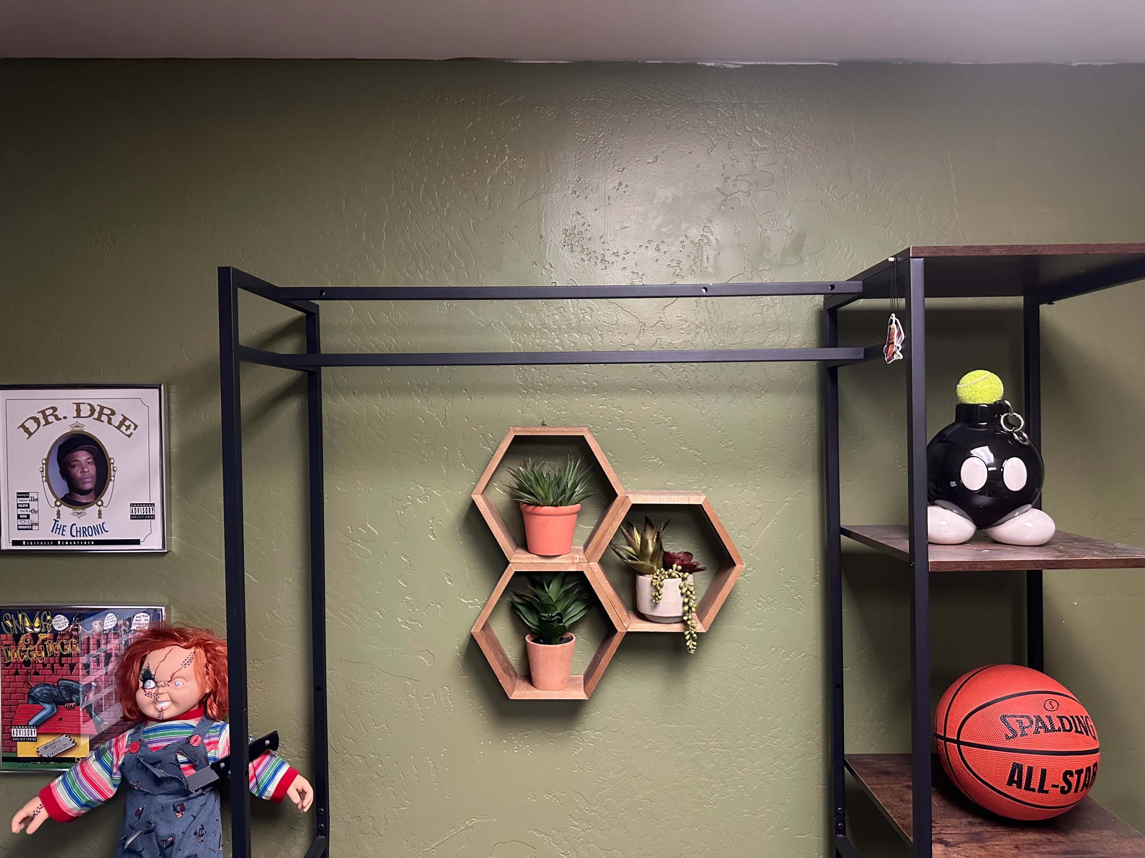 The image shows a wall painted green with a metal clothing rack, a hexagonal plant display, a shelf with a basketball, and two dolls, one resembling a famous horror character.