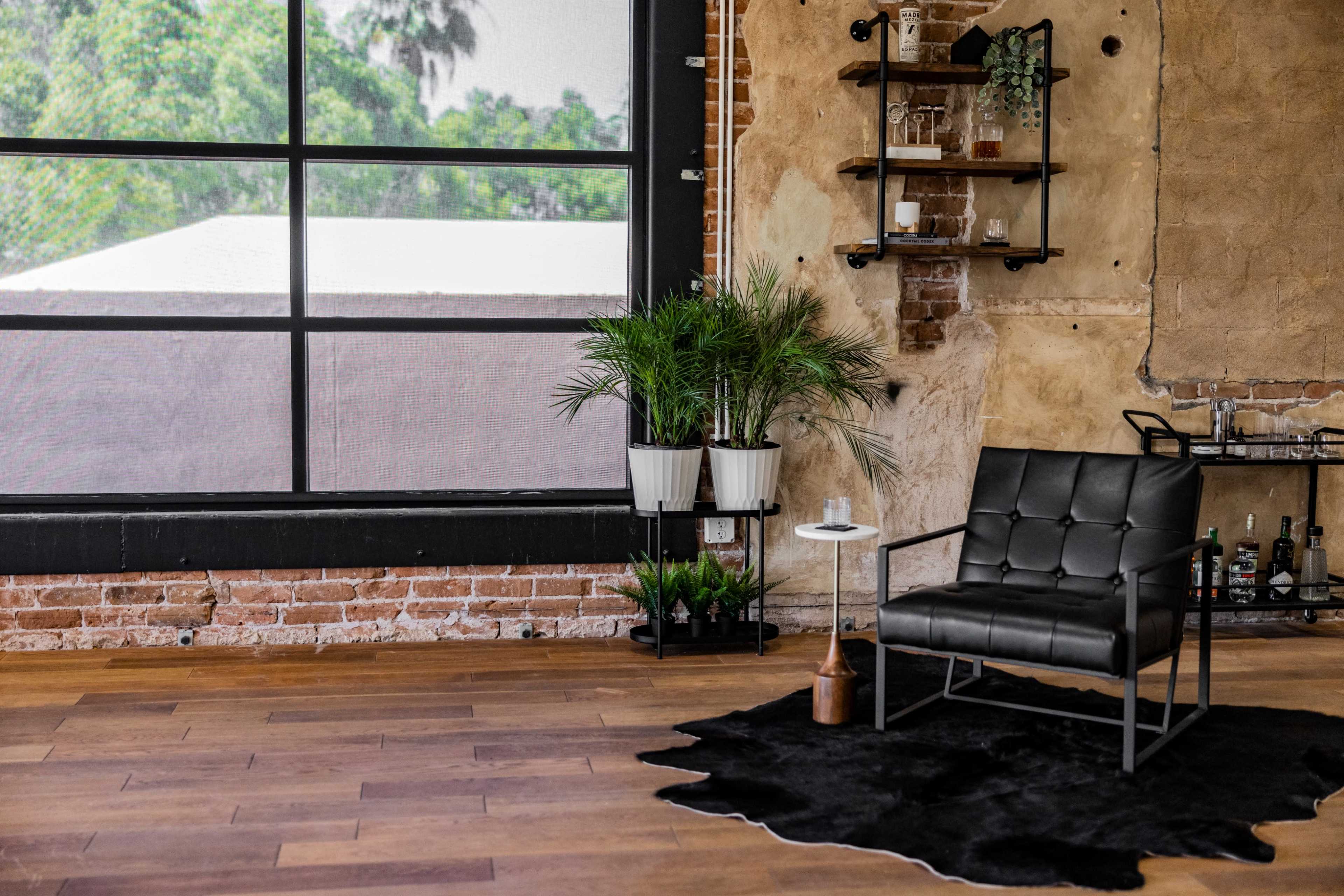 A black armchair sits beside a small table in a room with exposed brick walls and large windows overlooking greenery.