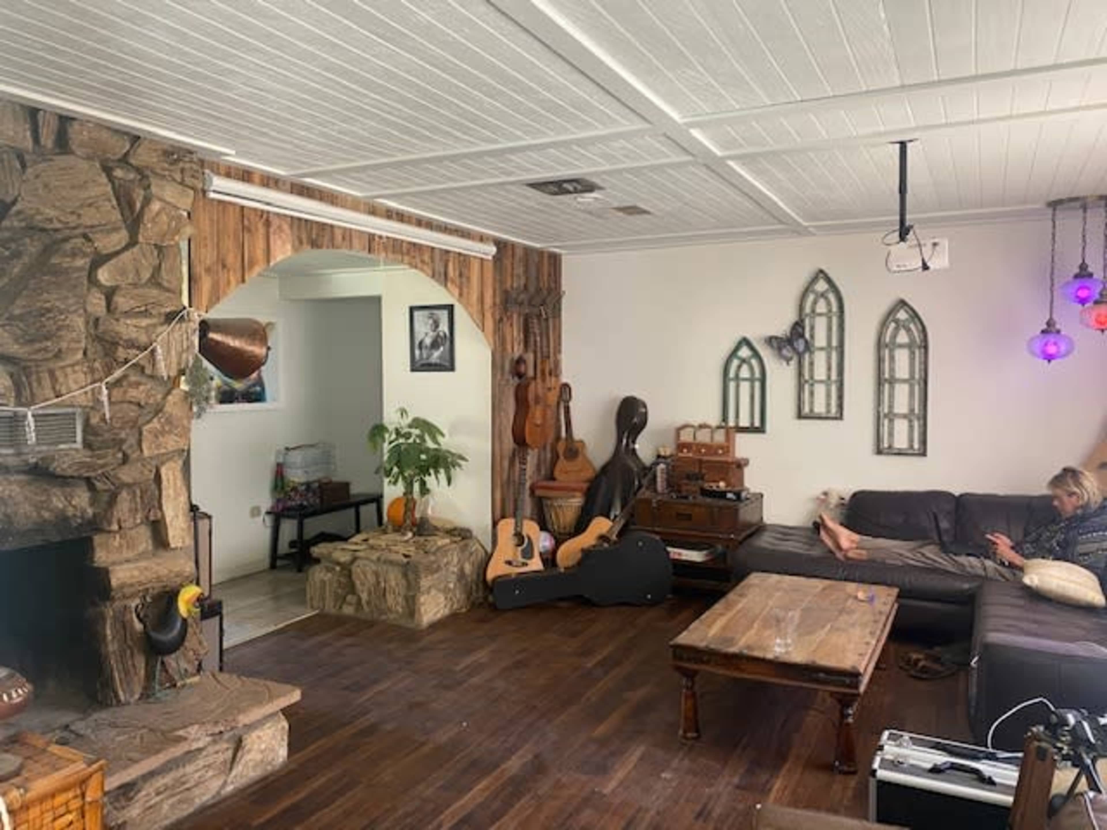 The image shows a living room featuring a stone fireplace, wooden accents, various musical instruments, and a person seated on a couch.
