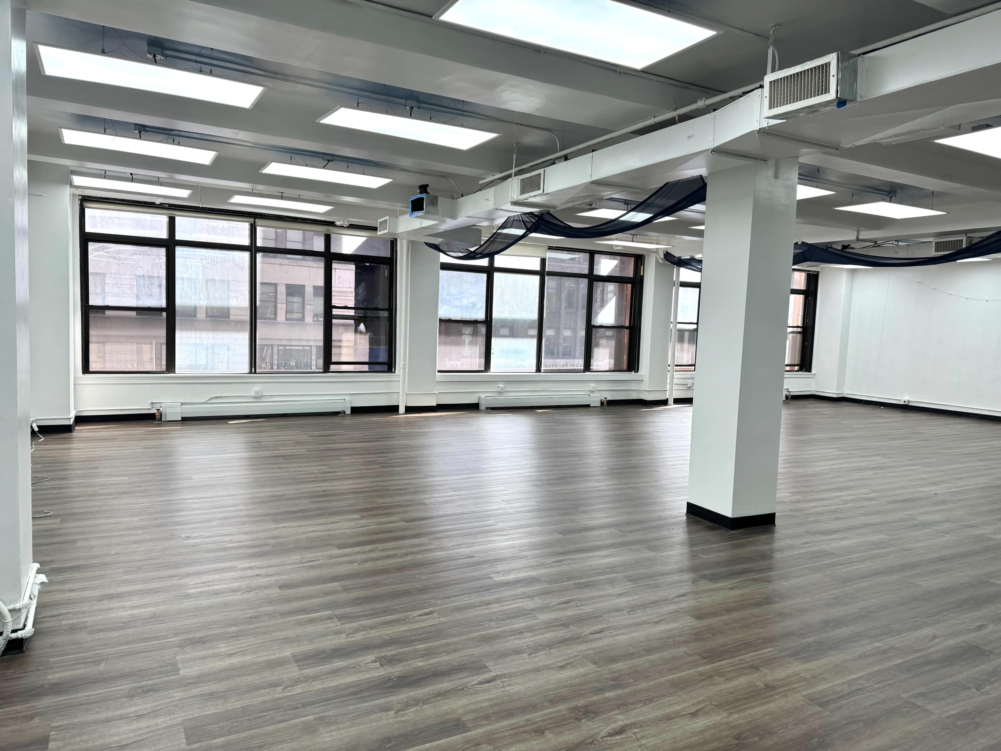 The image shows a spacious, empty room with large windows, white walls, and wooden flooring.