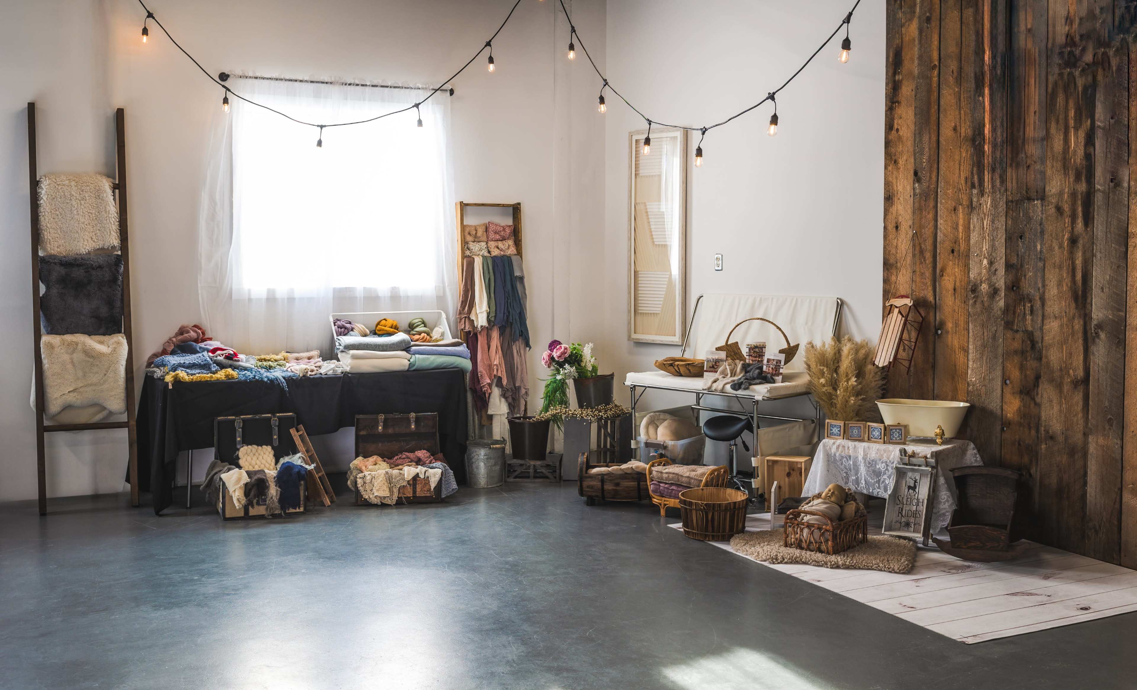 A brightly lit room features various textiles and baskets neatly arranged on tables and the floor, with string lights overhead and wooden walls in the background.