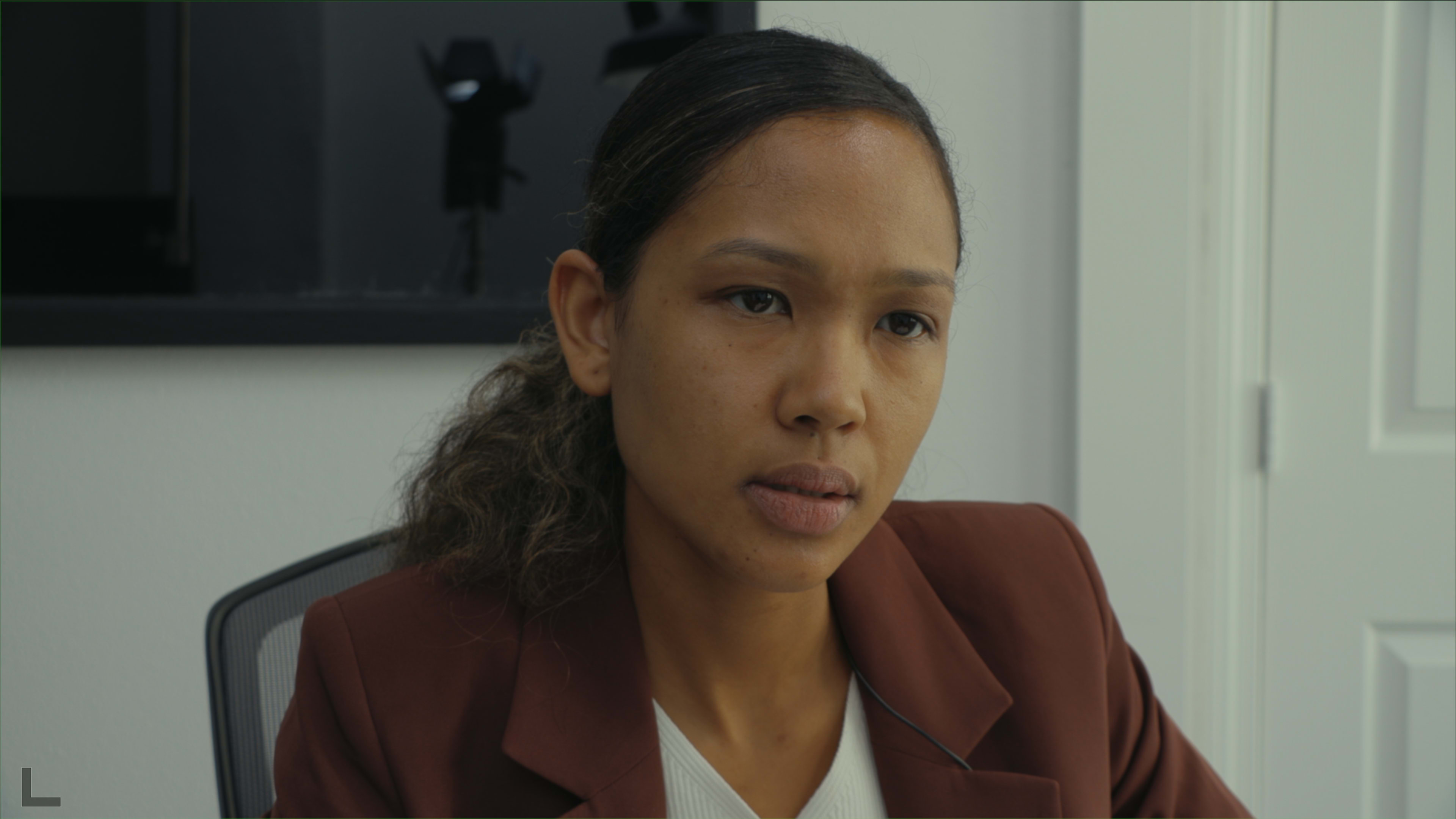 A woman with straight hair sits at a desk, wearing a brown blazer, with a focused expression.