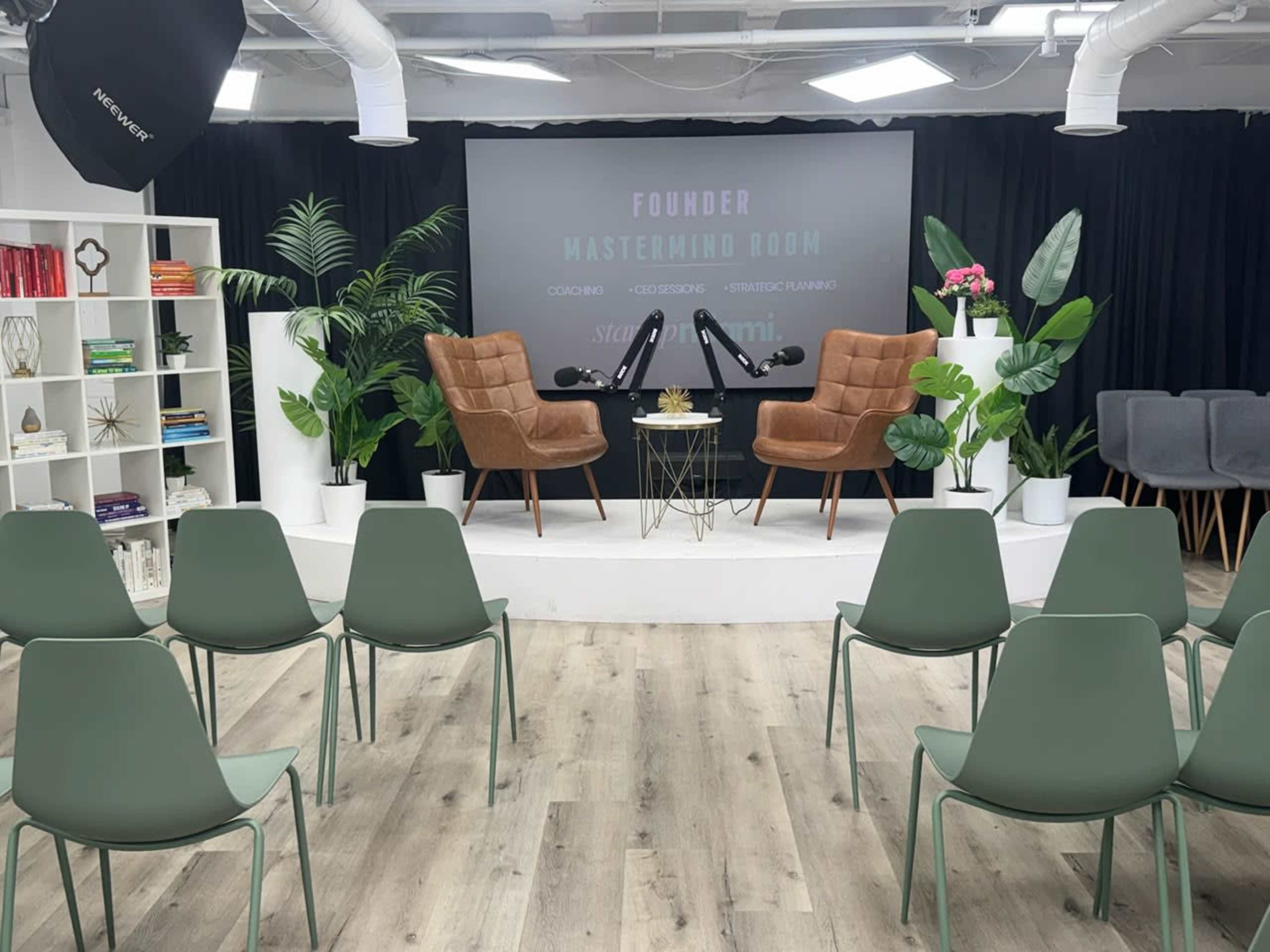 The image shows a modern seminar room with two brown chairs on a stage, flanked by potted plants, facing rows of green chairs, and featuring a backdrop for presentations.