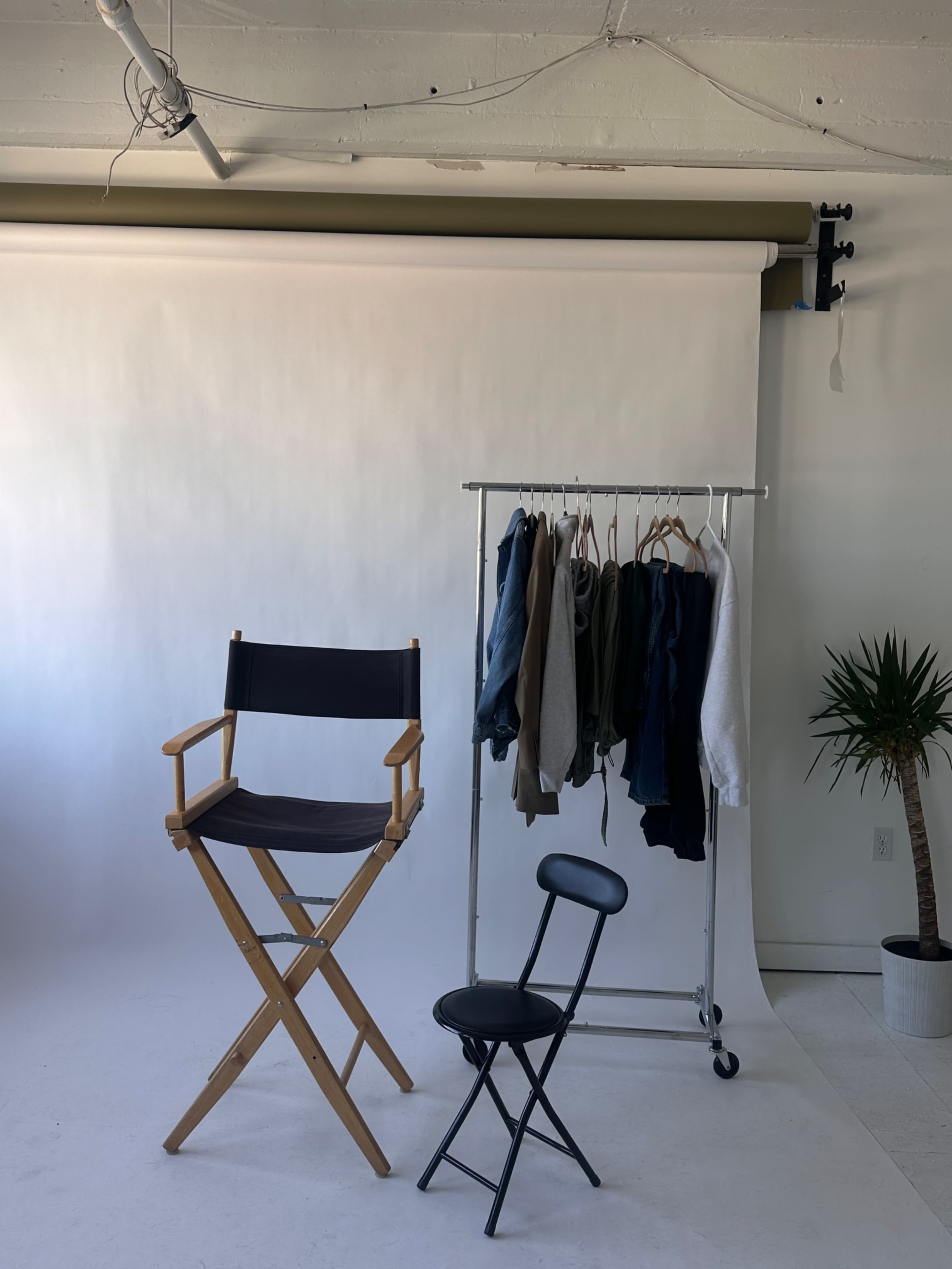 A wooden director's chair and a black chair are positioned next to a clothing rack holding various sweatshirts and jackets in a photo studio with a white backdrop.