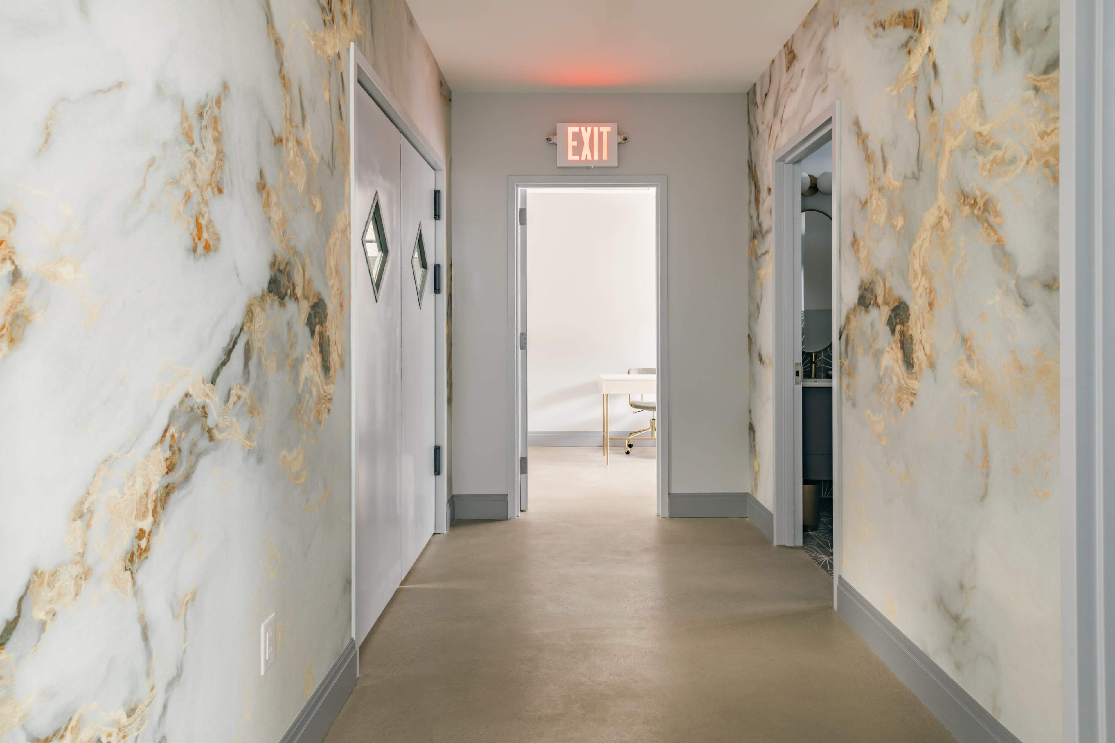 A corridor with textured walls, an exit sign, and an open door leading to a bright room with furniture.