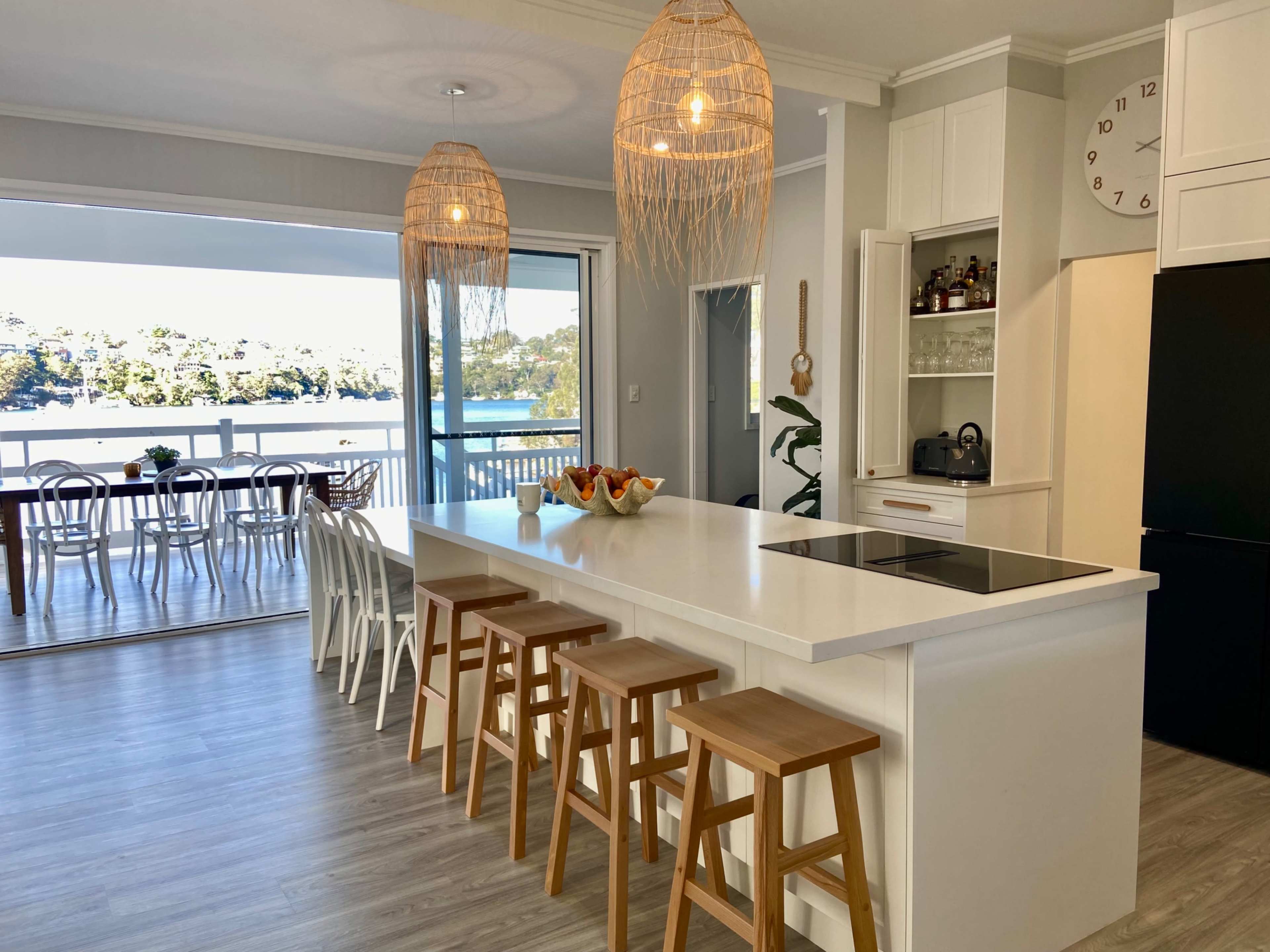 A modern kitchen features a central island with wooden stools and pendant lighting, overlooking a dining area and a scenic view of water and greenery.