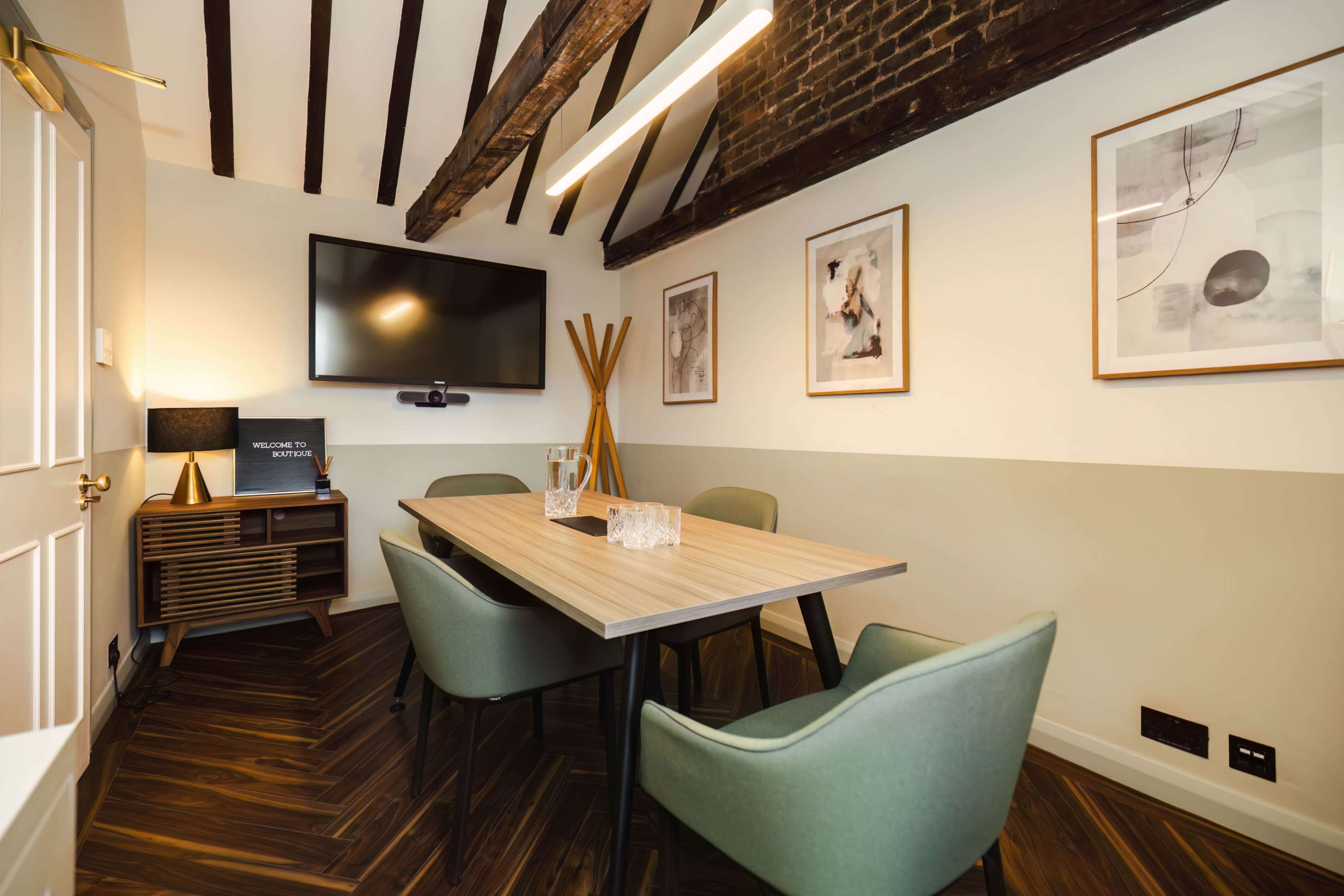 A conference room features a wooden table surrounded by green chairs, with a television mounted on the wall and framed artwork displayed.