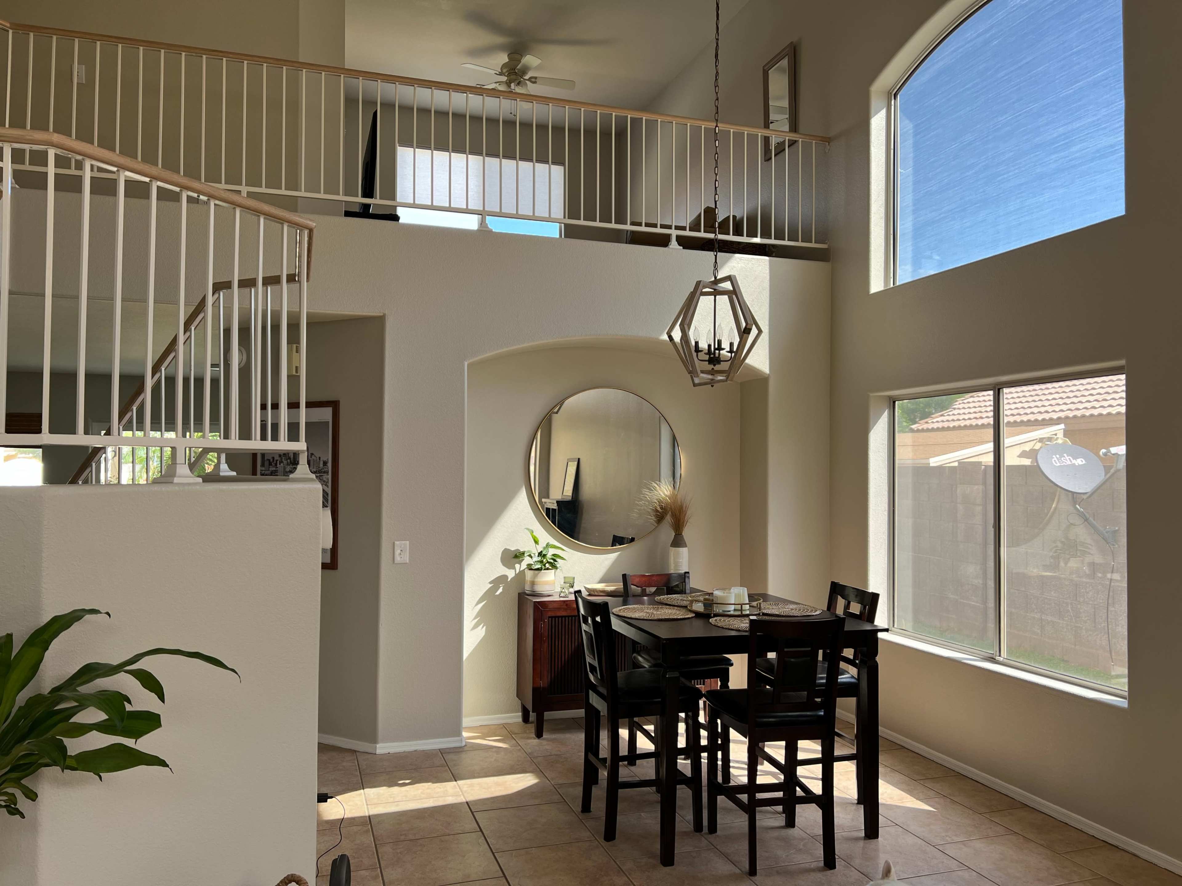 A spacious dining area with a round mirror and a black dining table set against a backdrop of large windows and a staircase.