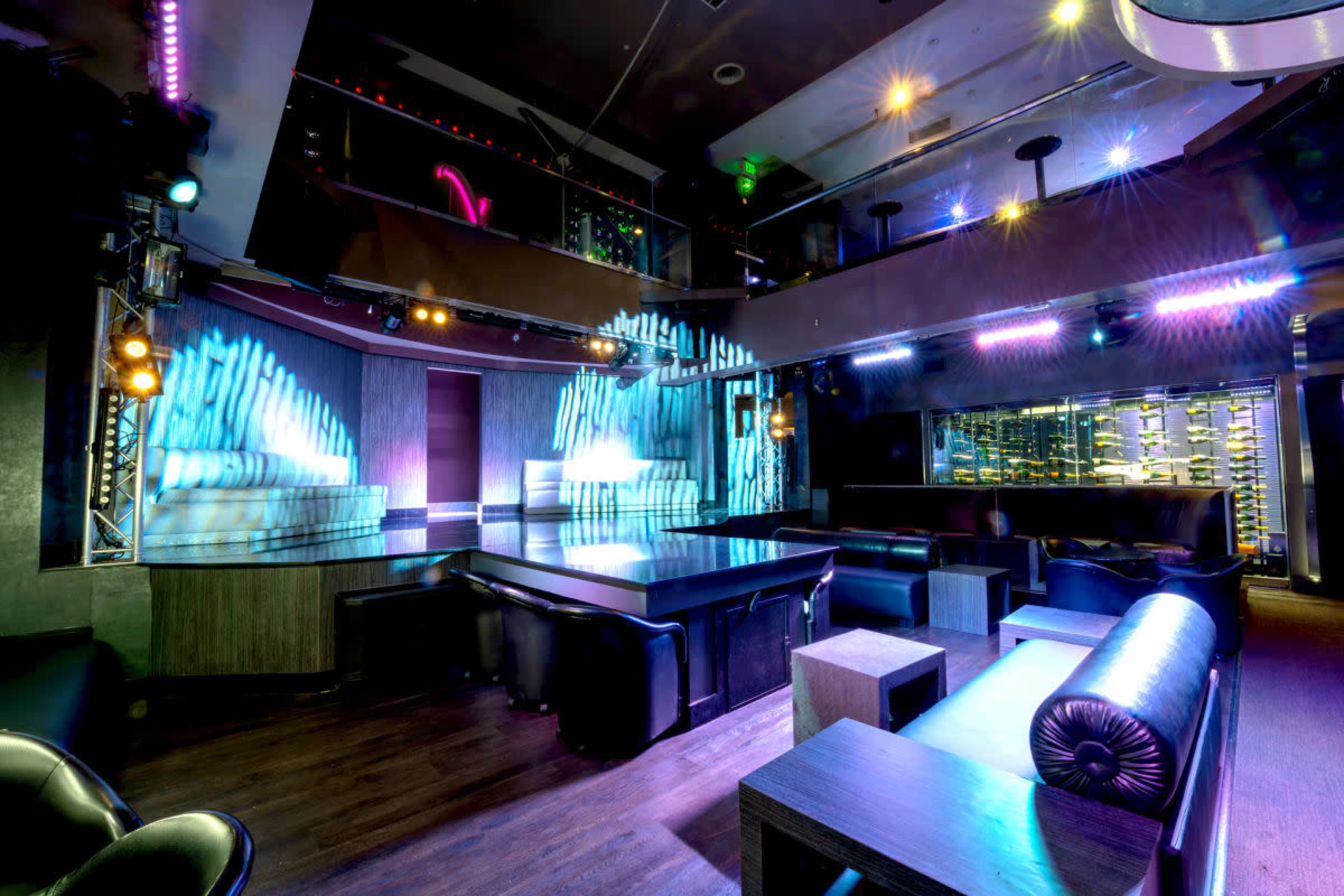 The image shows a modern nightclub interior with lounge seating, colorful lighting, and a stage area.