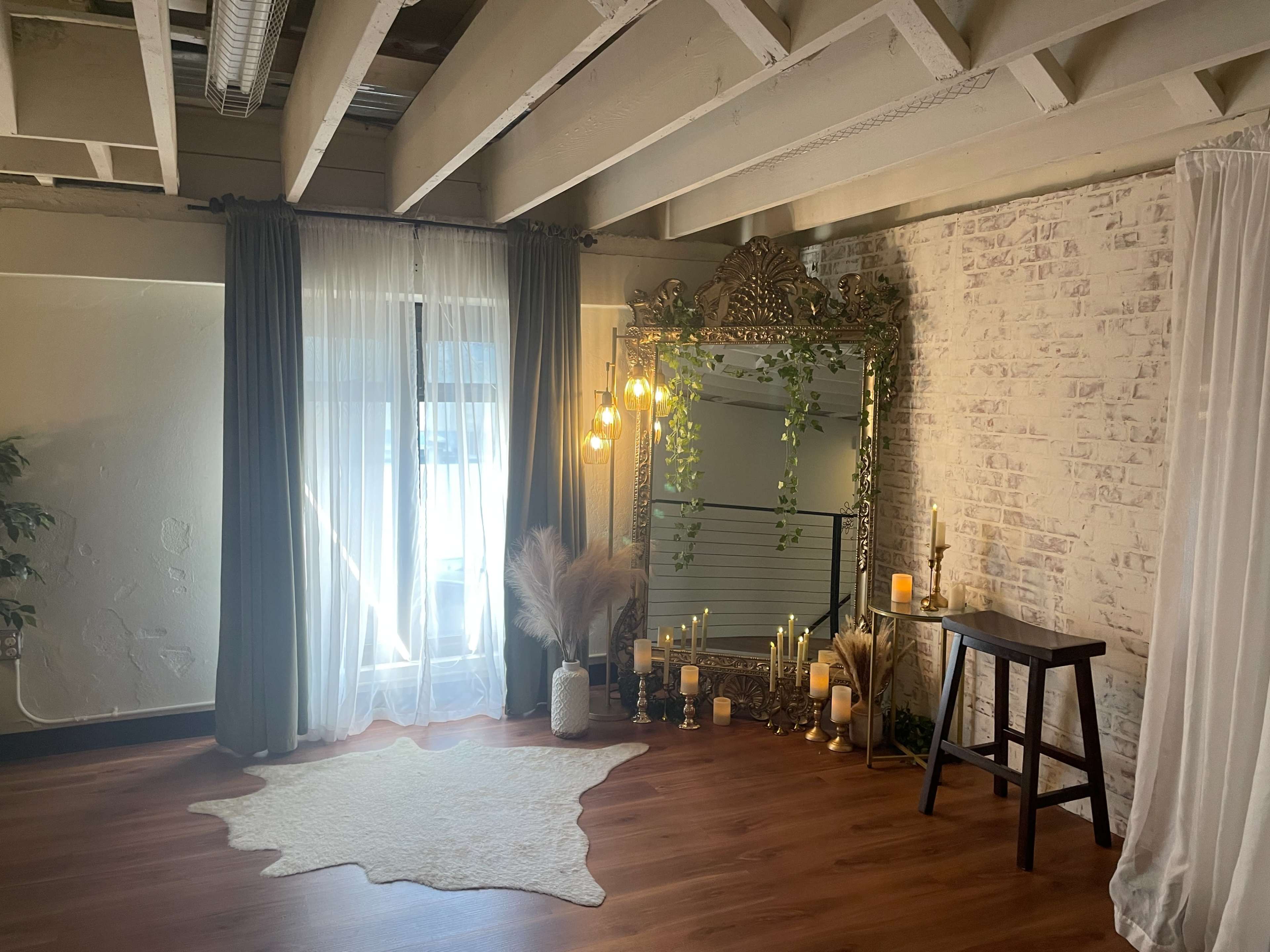 The image shows a well-lit room featuring a large ornate mirror, a cozy rug, decorative candles, and plants, with sheer curtains letting in natural light from a window.