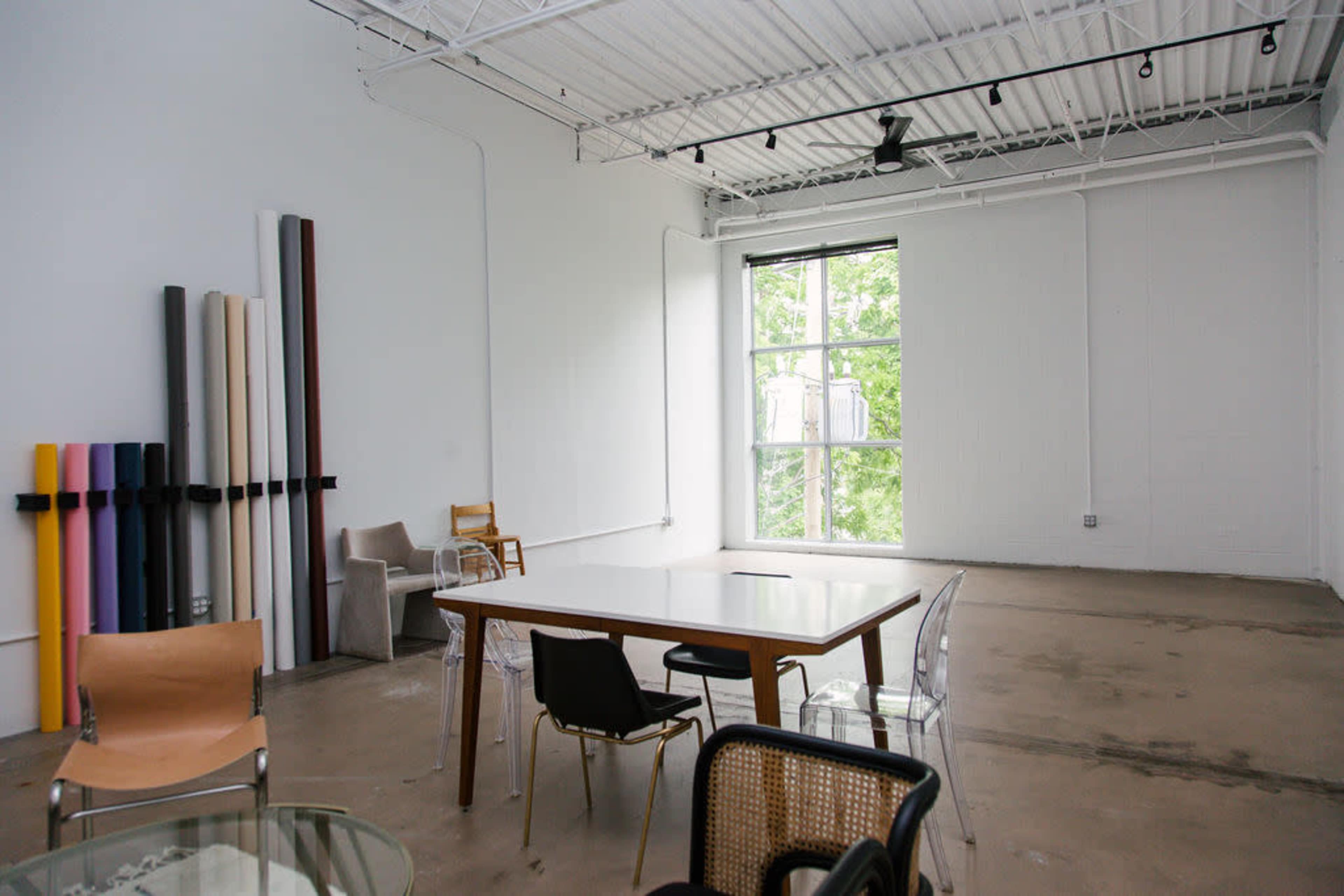 An empty studio space featuring a large table, several chairs, and rolls of material against the wall, with natural light coming through a large window.