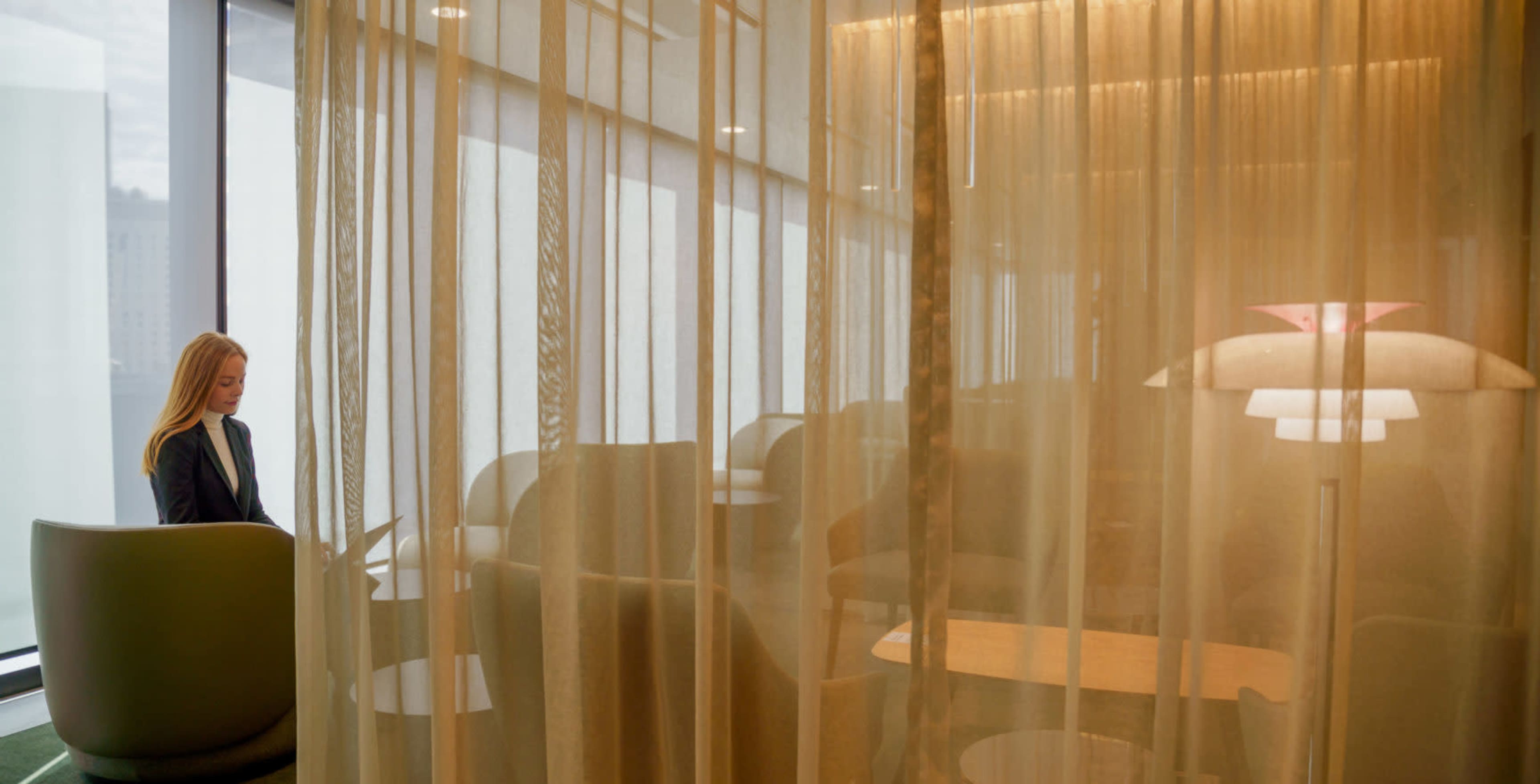 A woman with long hair sits in a modern, softly lit lounge area behind a sheer curtain.