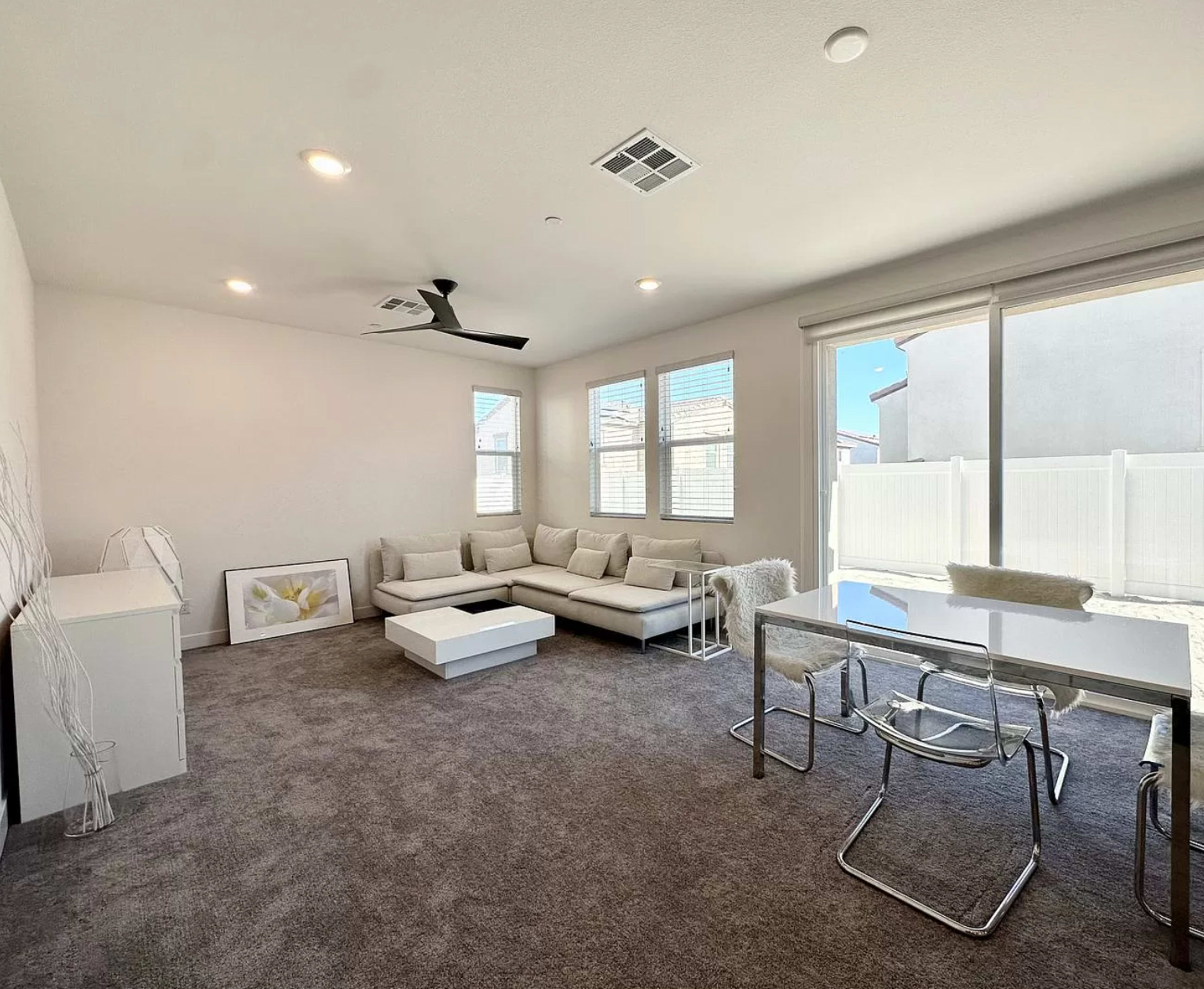 The image shows a modern living room with a sectional sofa, a coffee table, and a dining area featuring a glass table and metal chairs, all set against a backdrop of large windows.