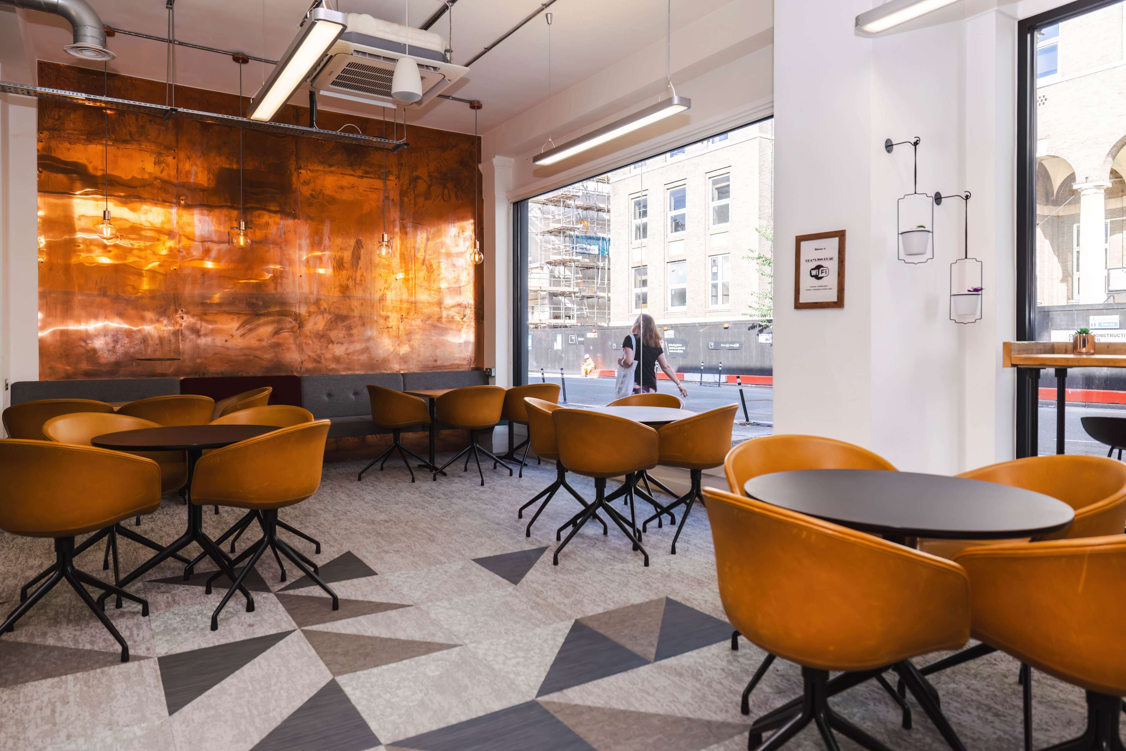 A modern cafe interior with wooden tables and brown chairs, featuring a large copper wall and large windows that open to a city view.