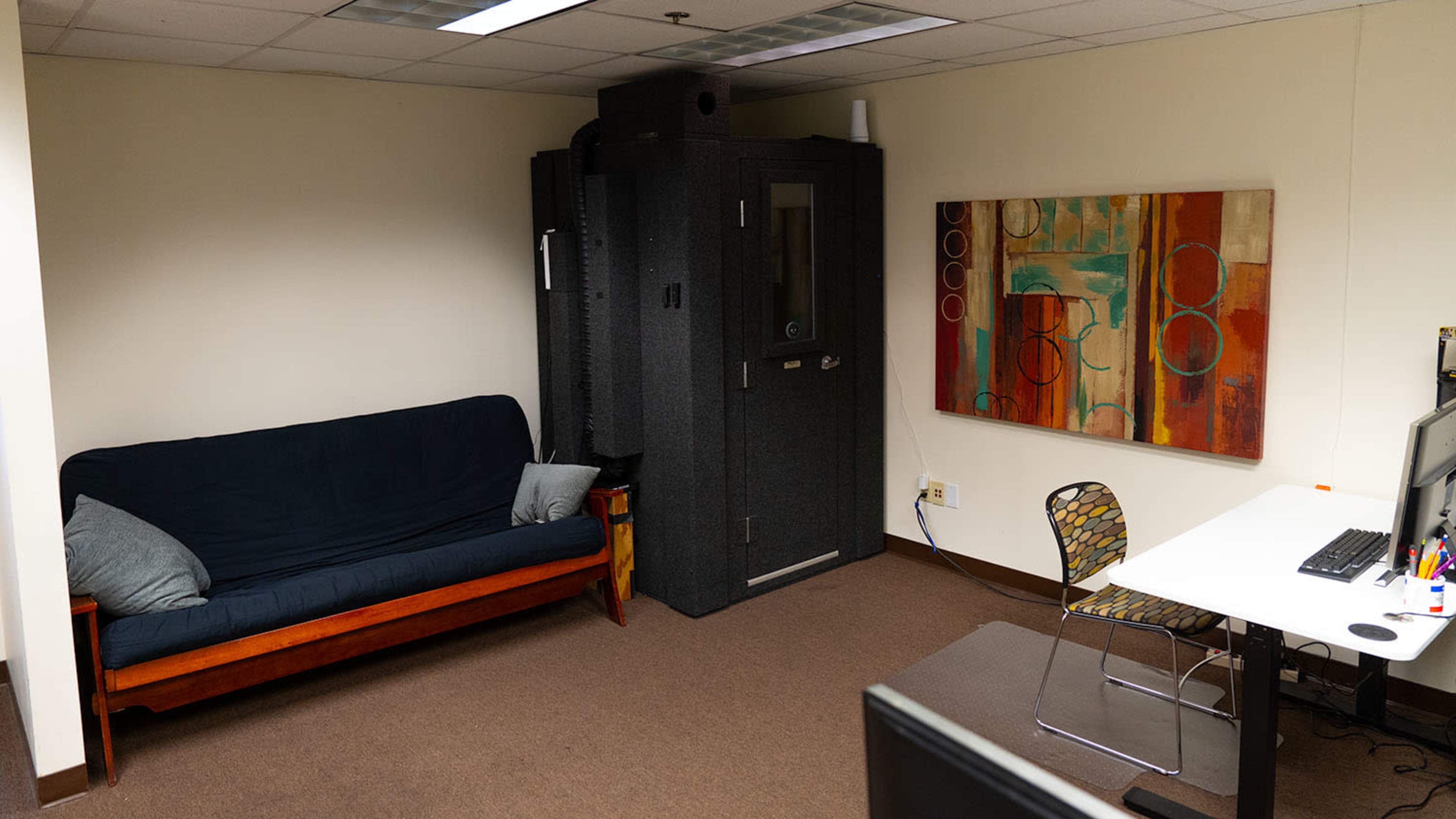 A small office space featuring a dark couch, a soundproof booth, an abstract painting on the wall, and a desk with a computer and a chair.