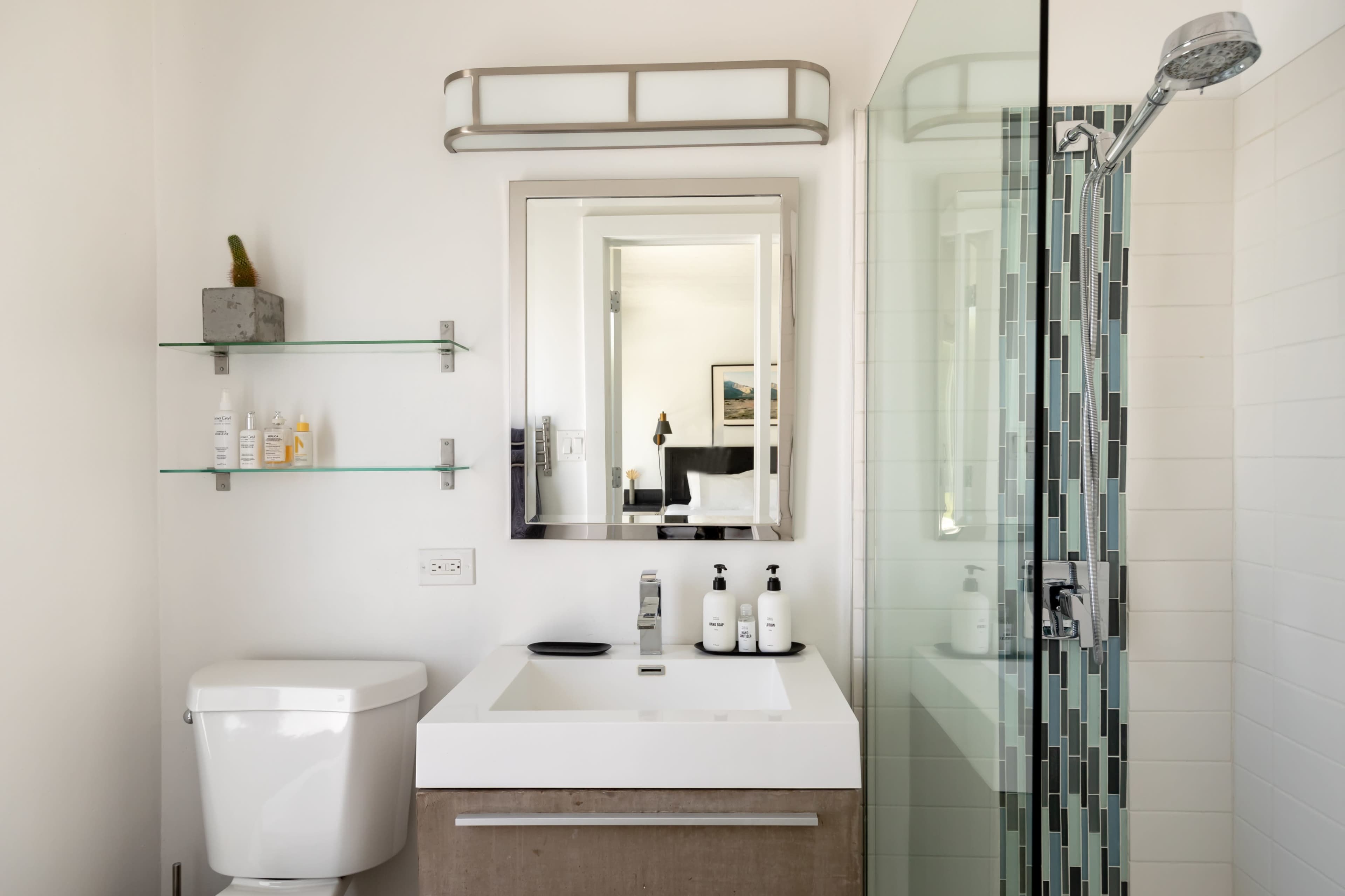 The image shows a modern bathroom featuring a toilet, a wall-mounted sink with a mirror above it, a glass shower enclosure, and open shelving with toiletries.