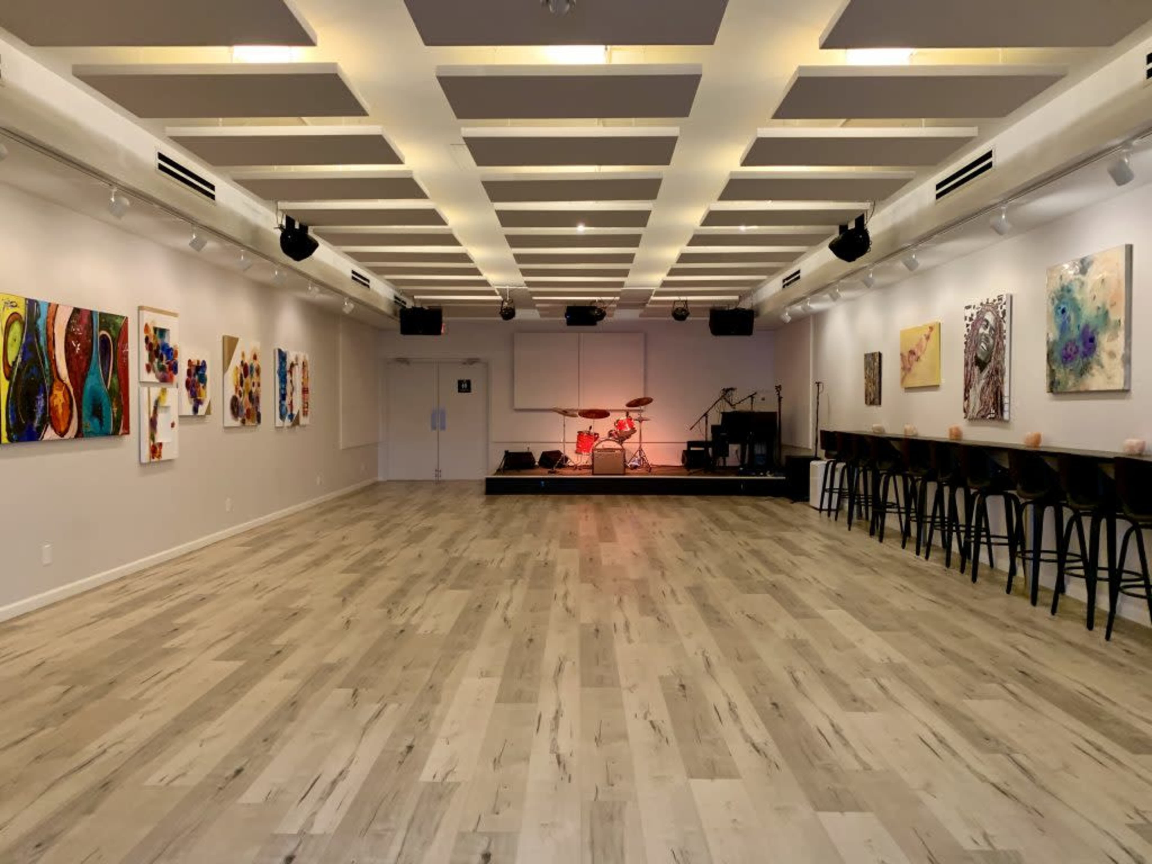 A spacious, well-lit performance venue with a polished wooden floor, featuring artwork on the walls and a stage set for a band at the far end.